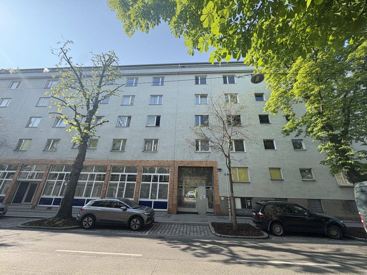 Straßenansicht eines Wohngebäudes mit Geschäften im Erdgeschoss und parkenden Autos, typische Stadtarchitektur.