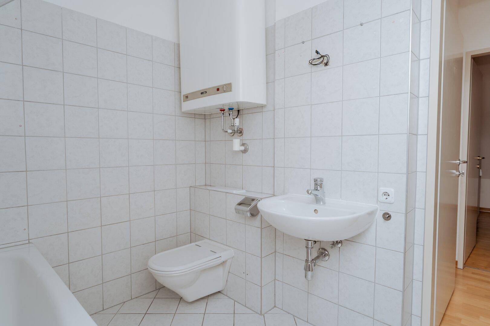 Funktionales Badezimmer mit weißen Fliesen, WC, Waschbecken und Warmwasserboiler.