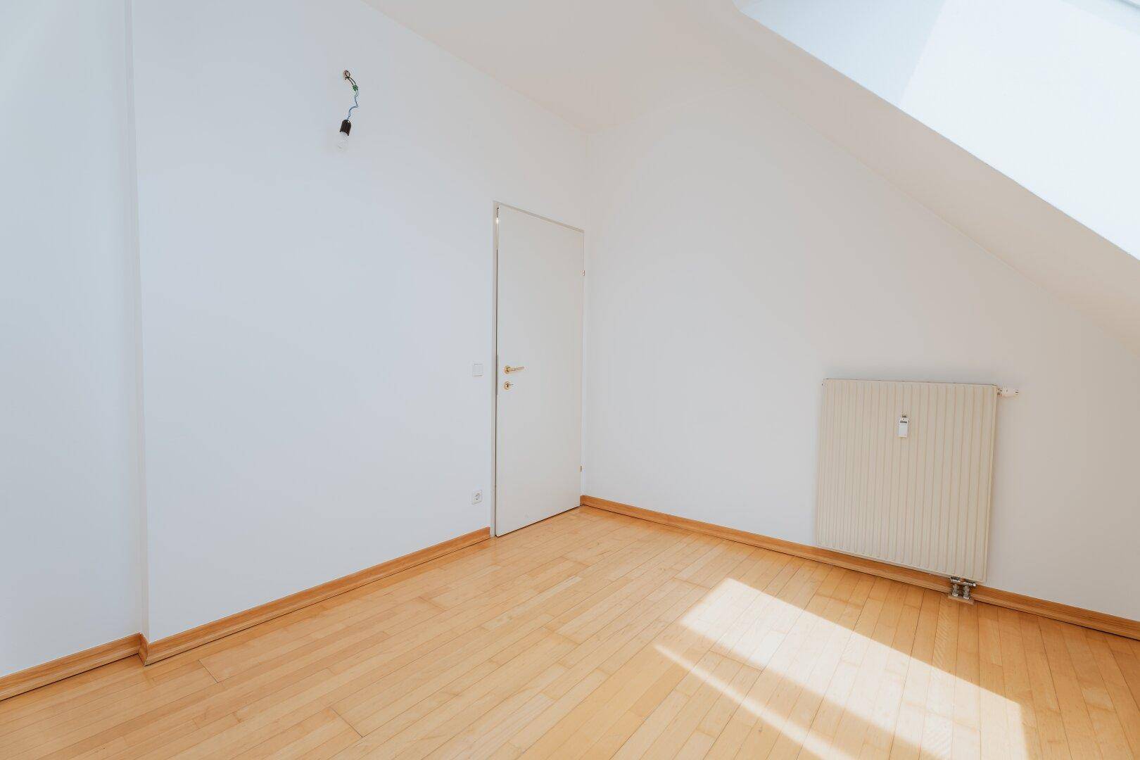 Schlafzimmer im Dachgeschoss mit Holzboden, weiß gestrichenen Wänden und Zugangstür.
