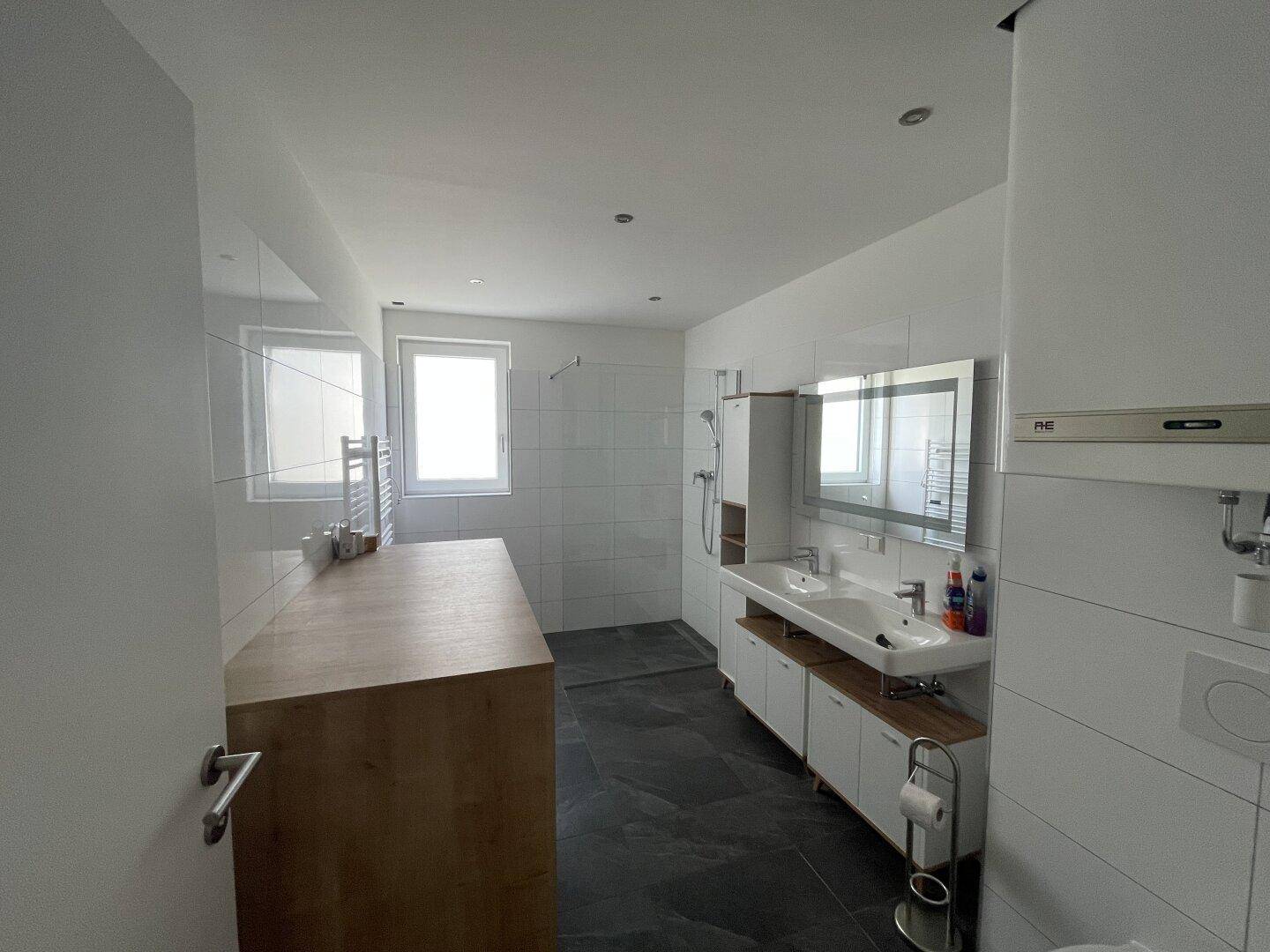 Modernes Badezimmer mit Doppelwaschbecken, großer Dusche, Fenster und dunklen Fliesen.