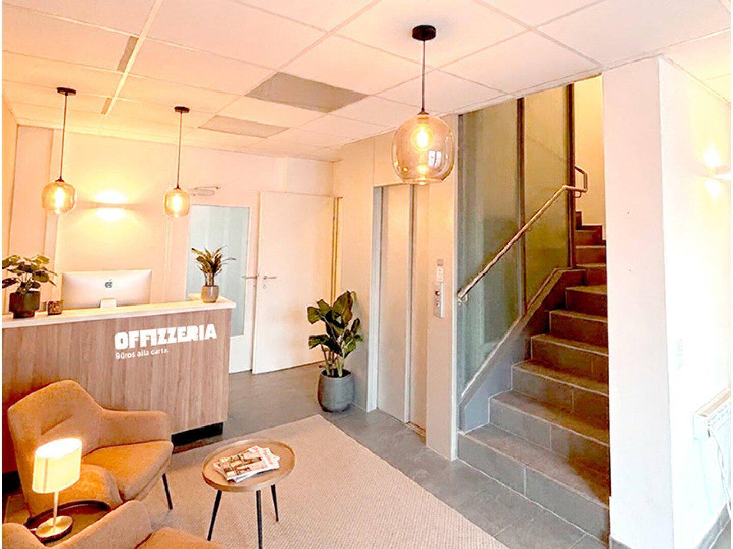 Empfangsbereich mit Rezeption, bequemen Sitzgelegenheiten, Aufzug und Treppe in einem modernen Gebäude.