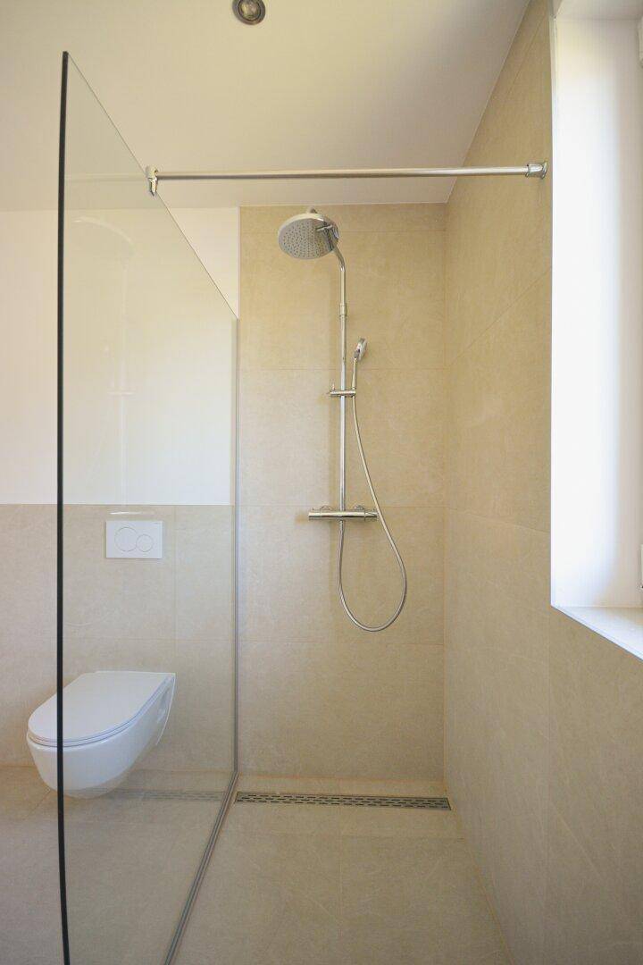 Modernes Badezimmer mit begehbarer Dusche, wandhängendem WC und hellen Fliesen, inklusive Fenster.
