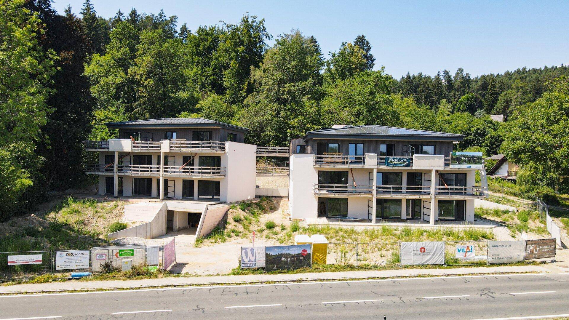 Zwei moderne Neubauten mit Balkonen und umgeben von Wald, nahe einer Straße gelegen.