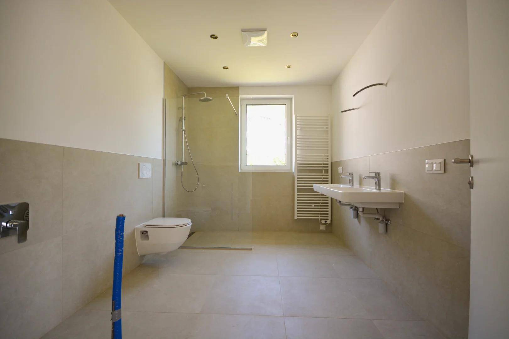 Modernes Badezimmer mit beigen Fliesen, bodengleicher Dusche, Doppelwaschbecken und Fenster für Tageslicht.