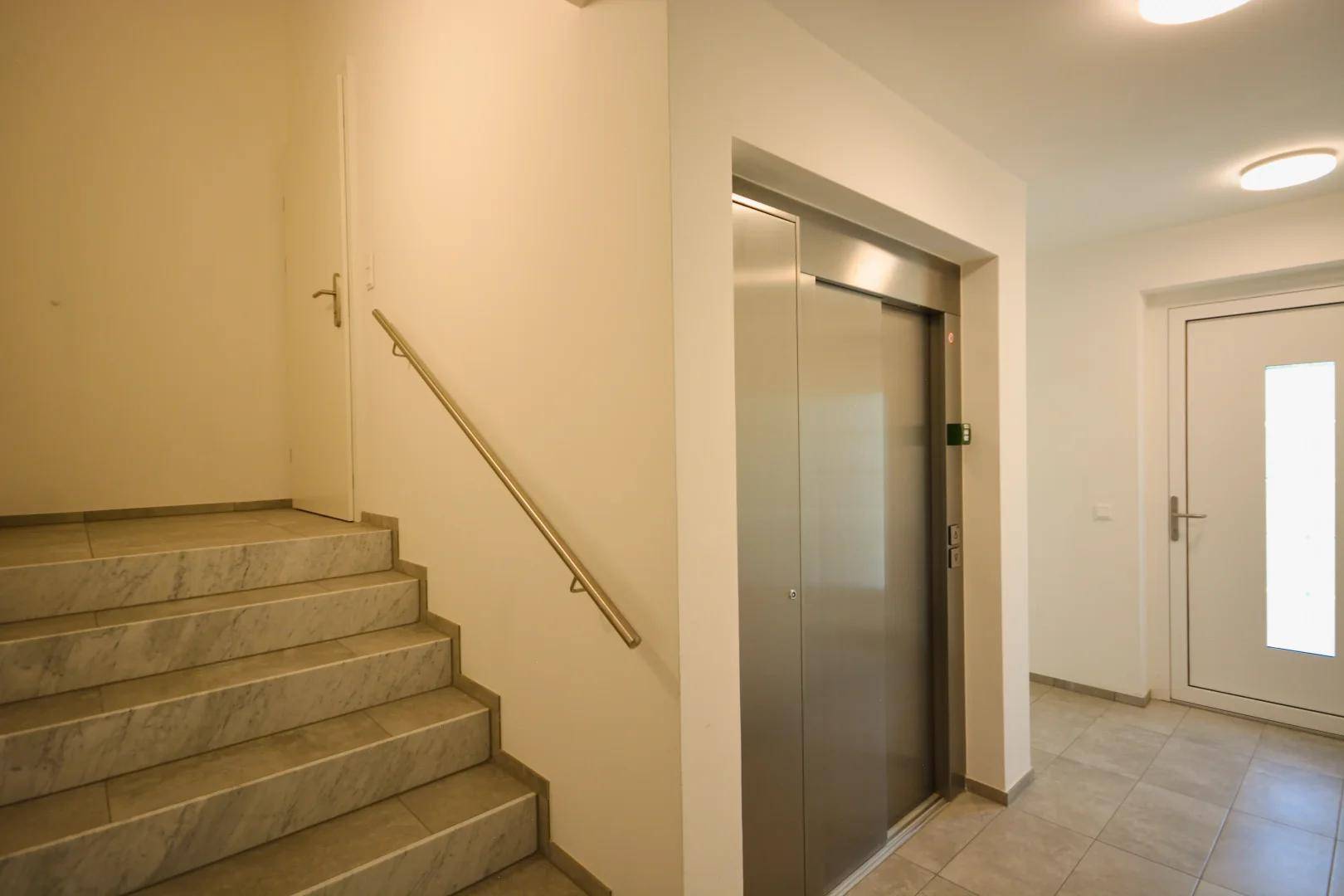 Heller Eingangsbereich mit Treppe und modernem Aufzug, der den Zugang zu den oberen Etagen erleichtert.