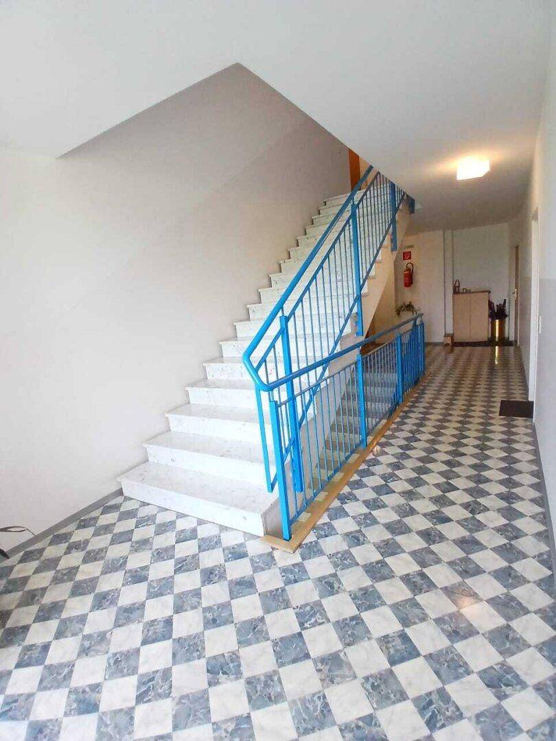 Treppenhaus mit blauen Geländern und auffälligem Schachbrettmuster-Fliesenboden im Gemeinschaftsbereich.