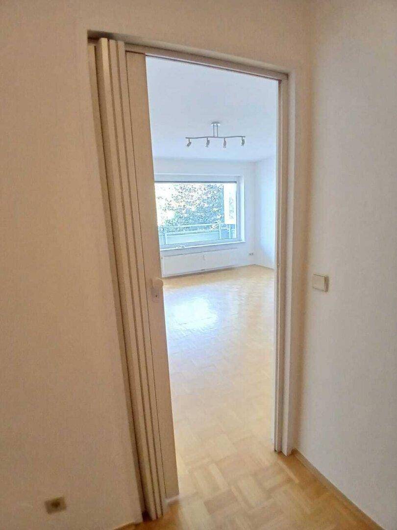 Blick durch eine offene Falttür in das helle Wohnzimmer mit Parkettboden und Balkonzugang.
