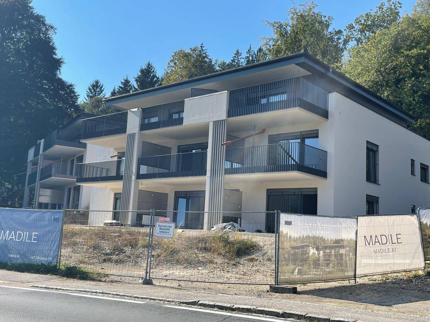 Modernes Mehrfamilienhaus im Bau mit mehreren Balkonen und Blick ins Grüne, umgeben von Bäumen.