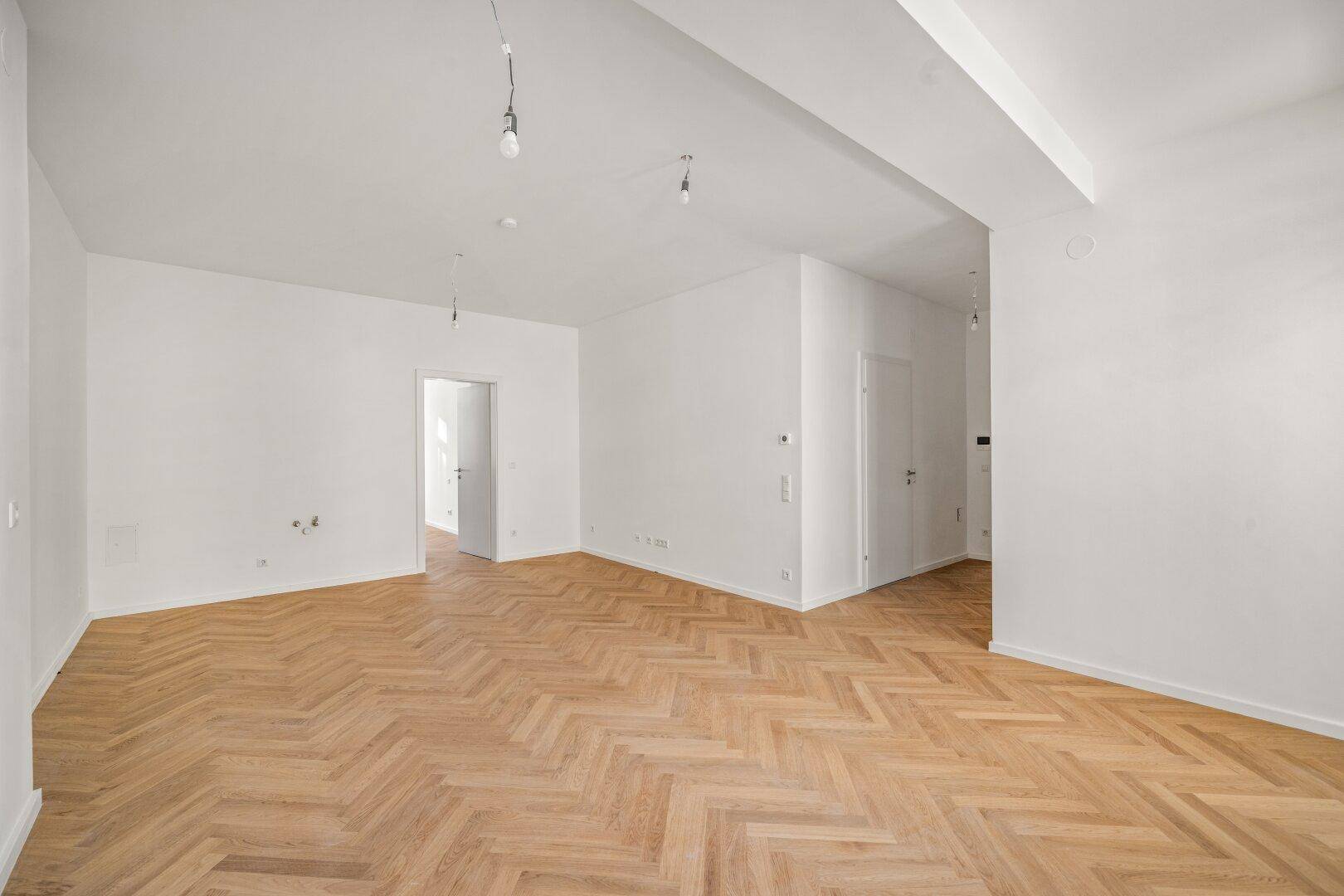Geräumiges, leeres Wohnzimmer mit hellem Fischgrätparkett und weißen Wänden, bereit zur Einrichtung.