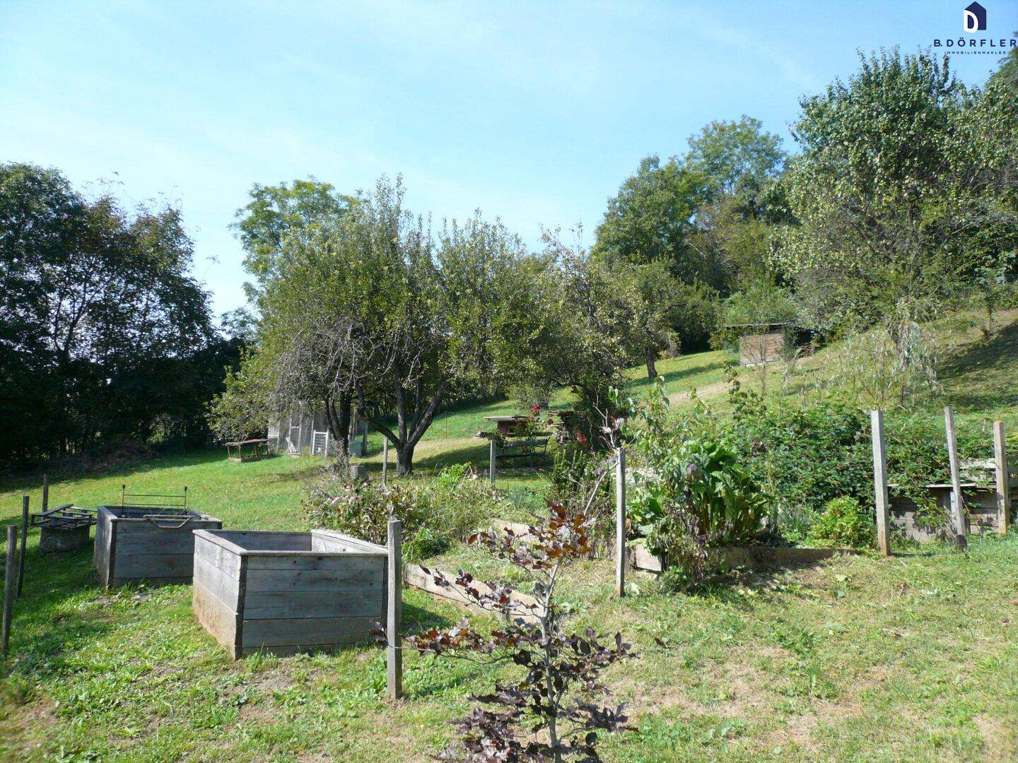 Naturbelassener Gartenbereich mit Hochbeeten und verschiedenen Bäumen in hügeliger Landschaft.