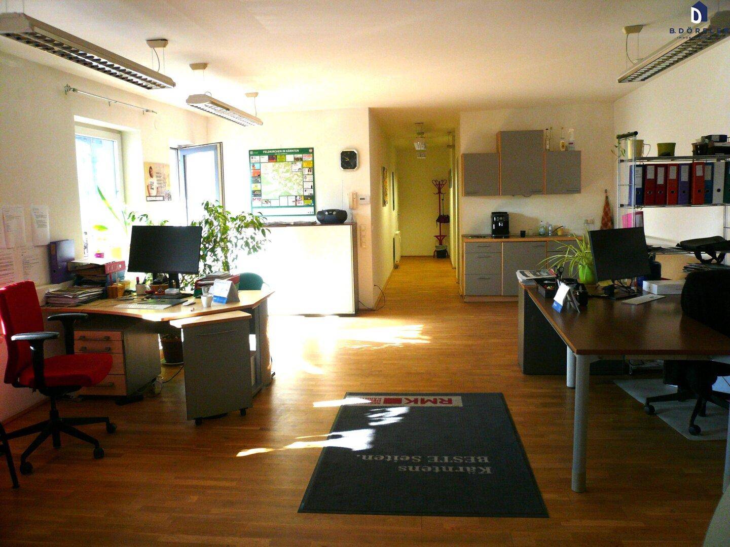 Helles und geräumiges Büro mit mehreren Arbeitsplätzen, Holzfußboden und viel Tageslicht durch große Fenster.