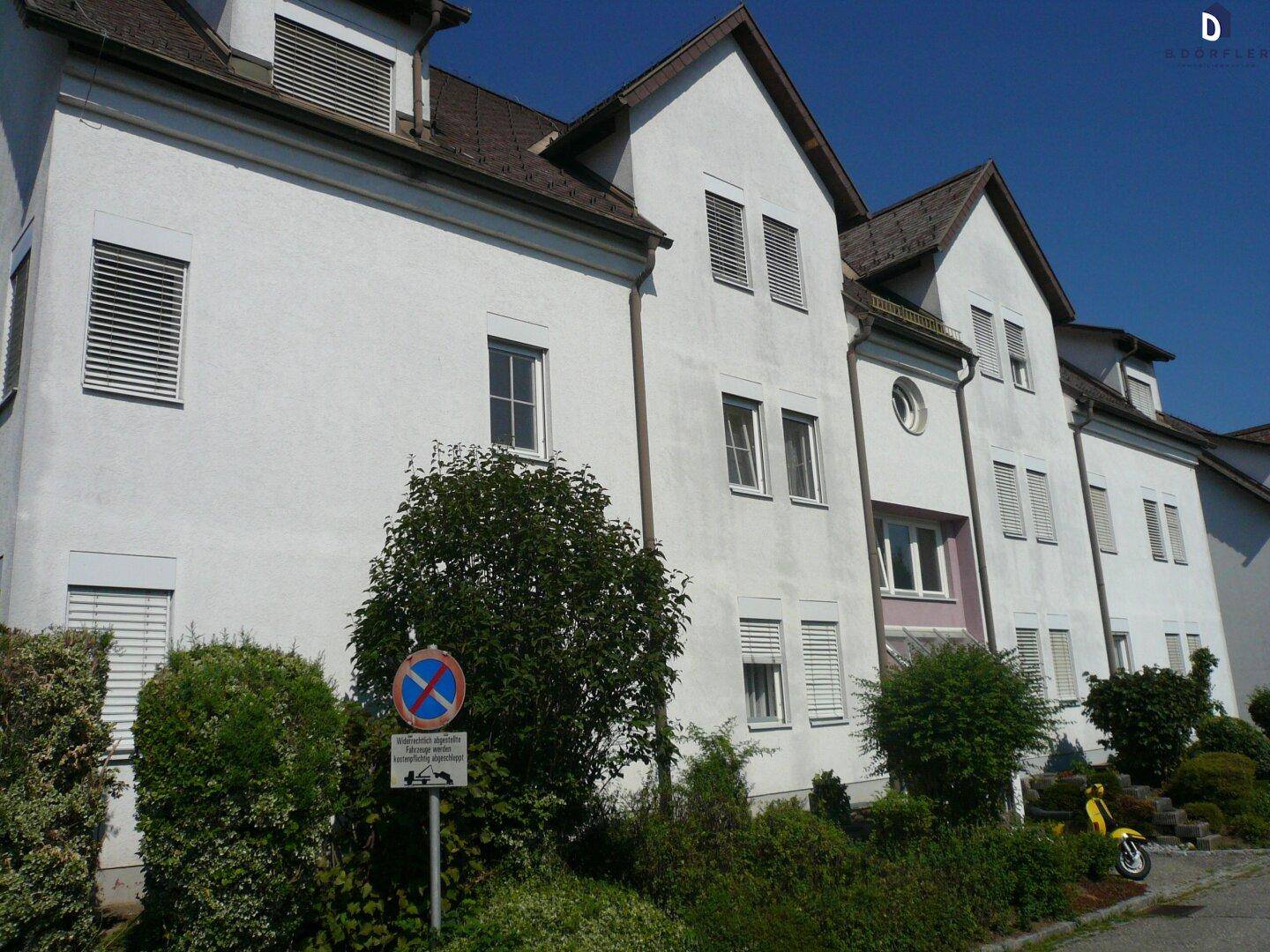 Weiße Wohngebäude mit mehreren Fenstern und Rollläden, umgeben von gepflegten Hecken.
