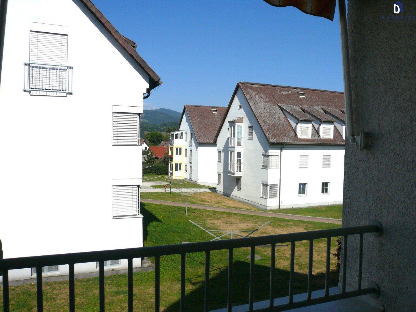 Blick von einem Balkon auf weitere Wohngebäude, grüne Rasenflächen und eine Berglandschaft.