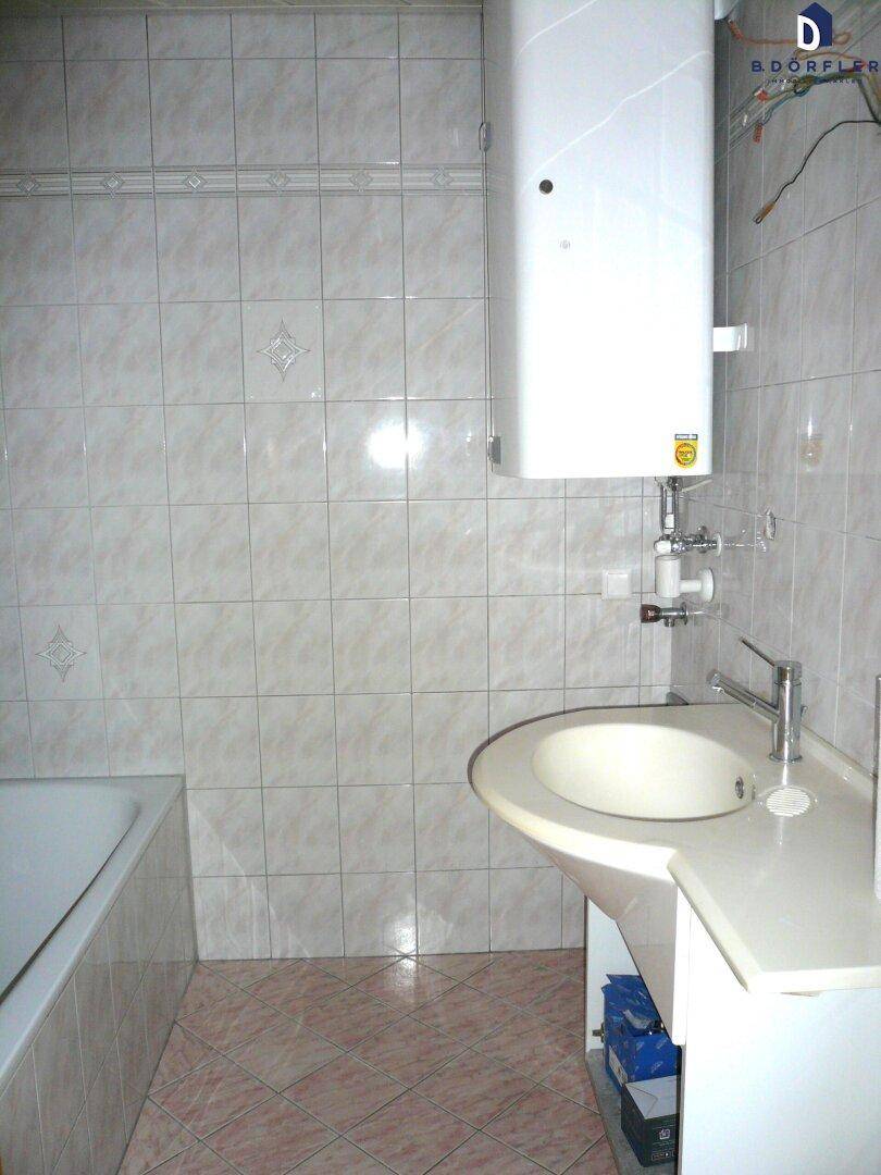 Badezimmer mit hellen Fliesen, Waschbecken, Badewanne und einem Warmwasserboiler.