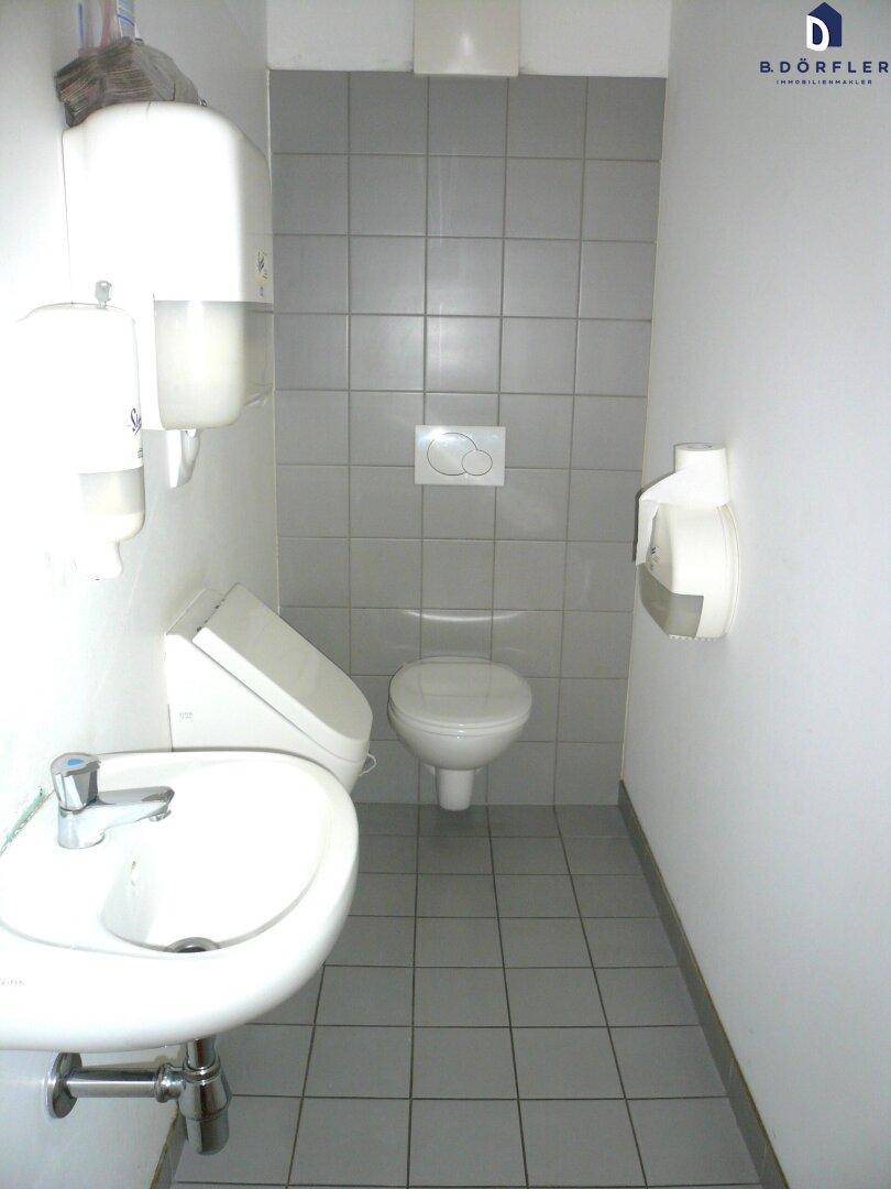 Kleine Toilette mit grau gefliestem Boden und Wänden, ausgestattet mit WC, Urinal und Waschbecken.