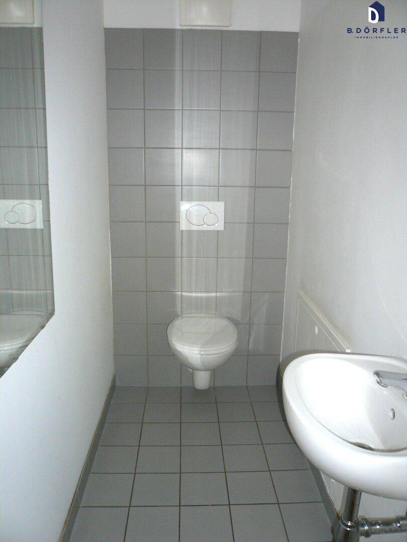 Saubere Toilette mit grauem Fliesenboden, Wand-WC und kleinem Waschbecken.
