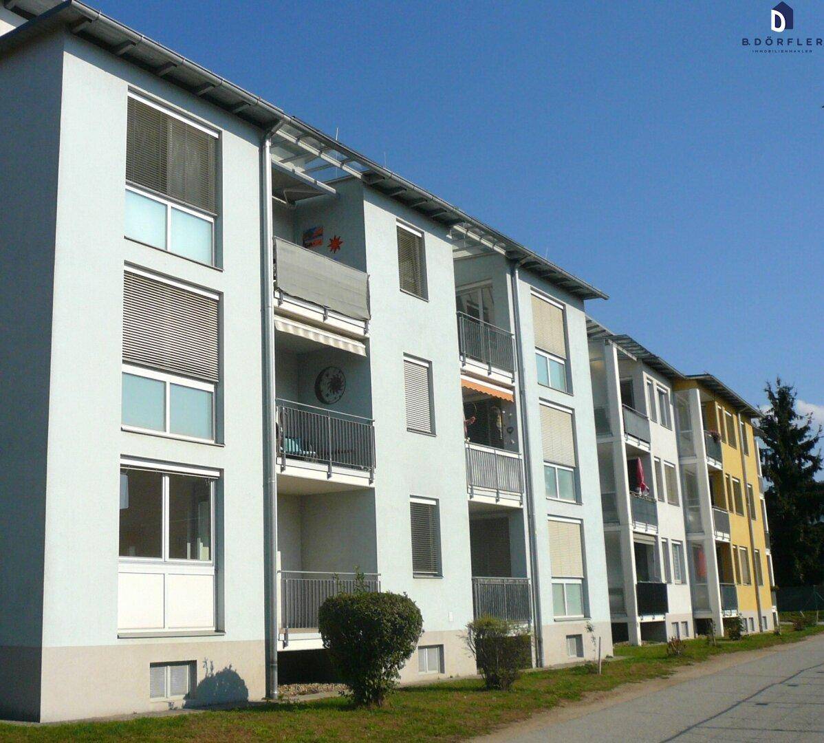 Mehrfamilienhaus mit mehreren Balkonen und Jalousien unter blauem Himmel, gut gepflegt.