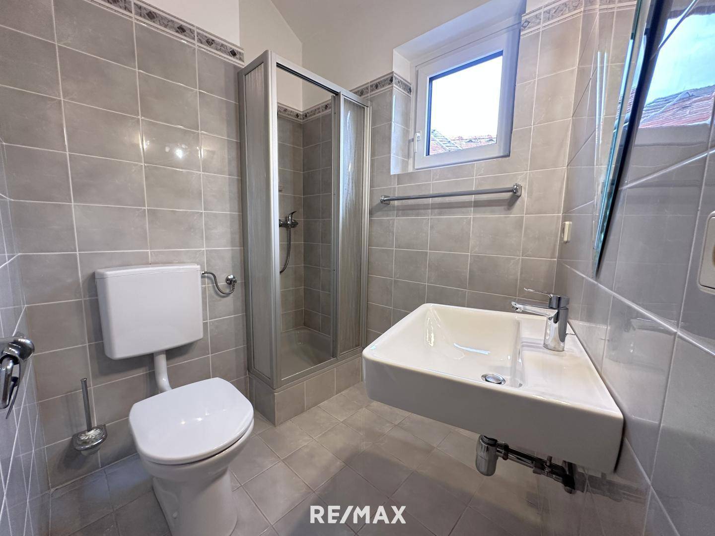 Modernes Badezimmer mit grauen Fliesen, Duschkabine und Fenster für Tageslicht.