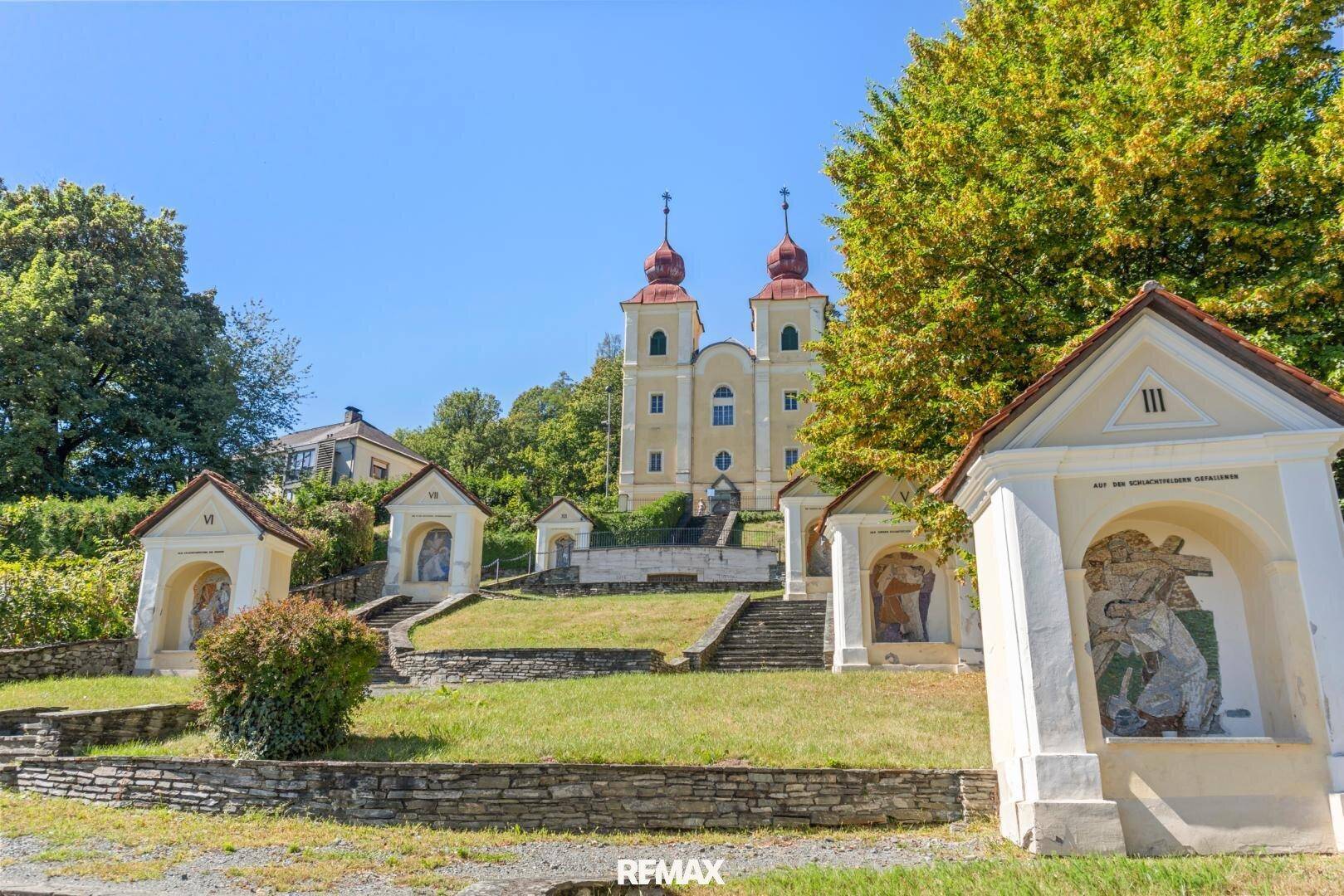 Historische Kirche mit zwei Türmen und Kreuzwegstationen in grüner Umgebung.