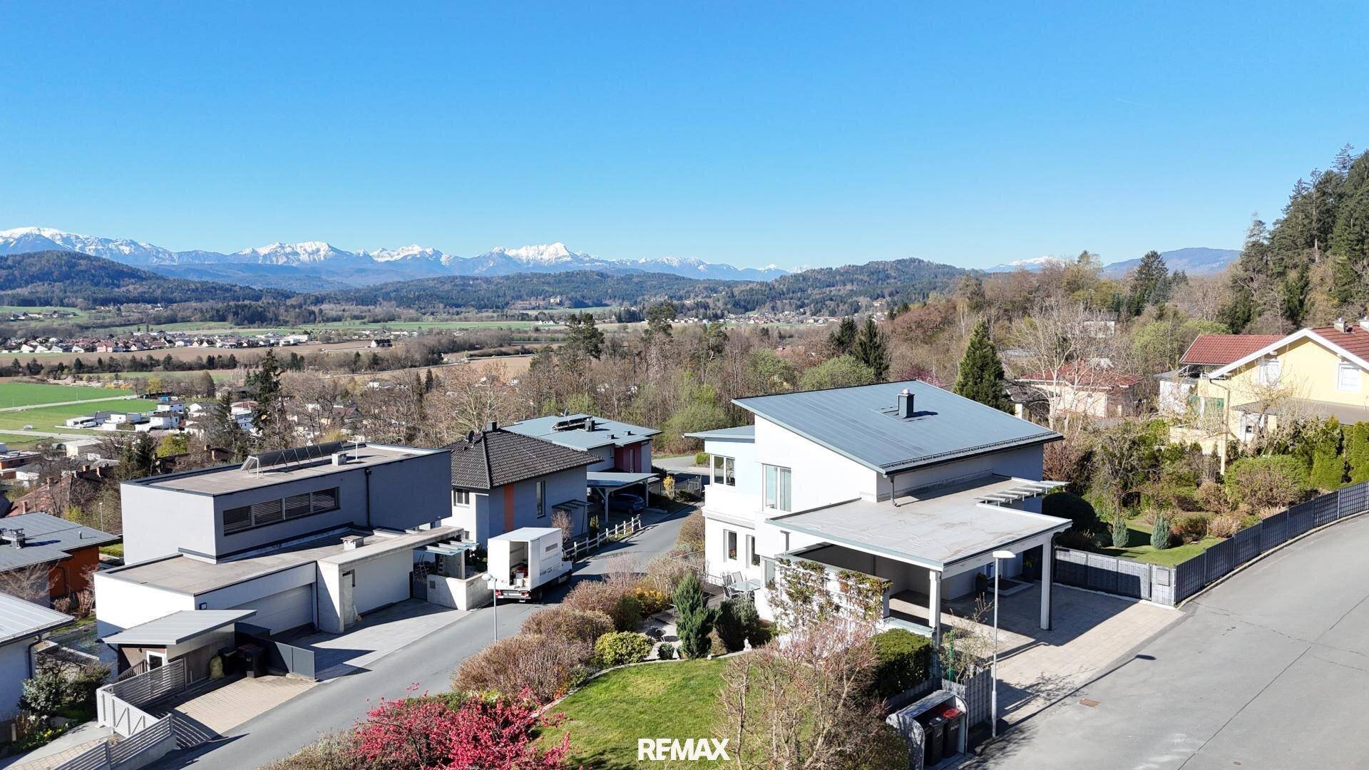Panoramablick auf eine moderne Wohnsiedlung mit Häusern, grünen Feldern und majestätischen Bergen im Hintergrund.