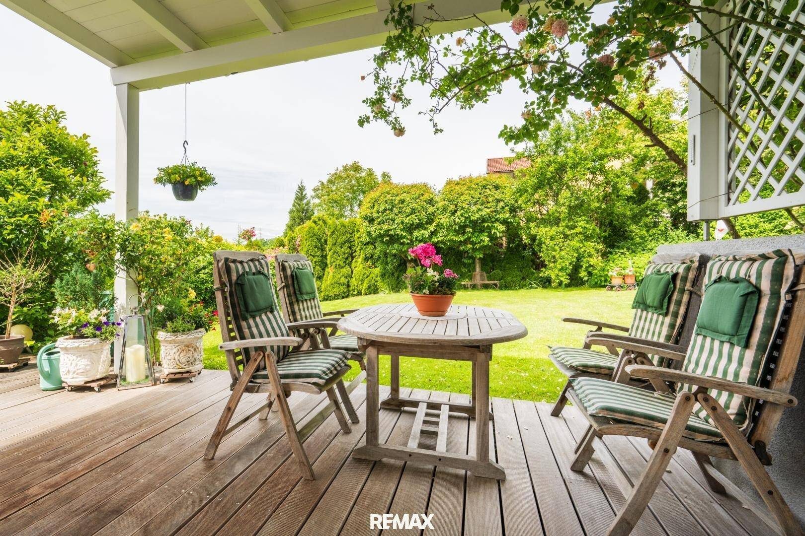 Überdachte Holzterrasse mit Gartenmöbeln, umgeben von einem üppigen, grünen Garten.