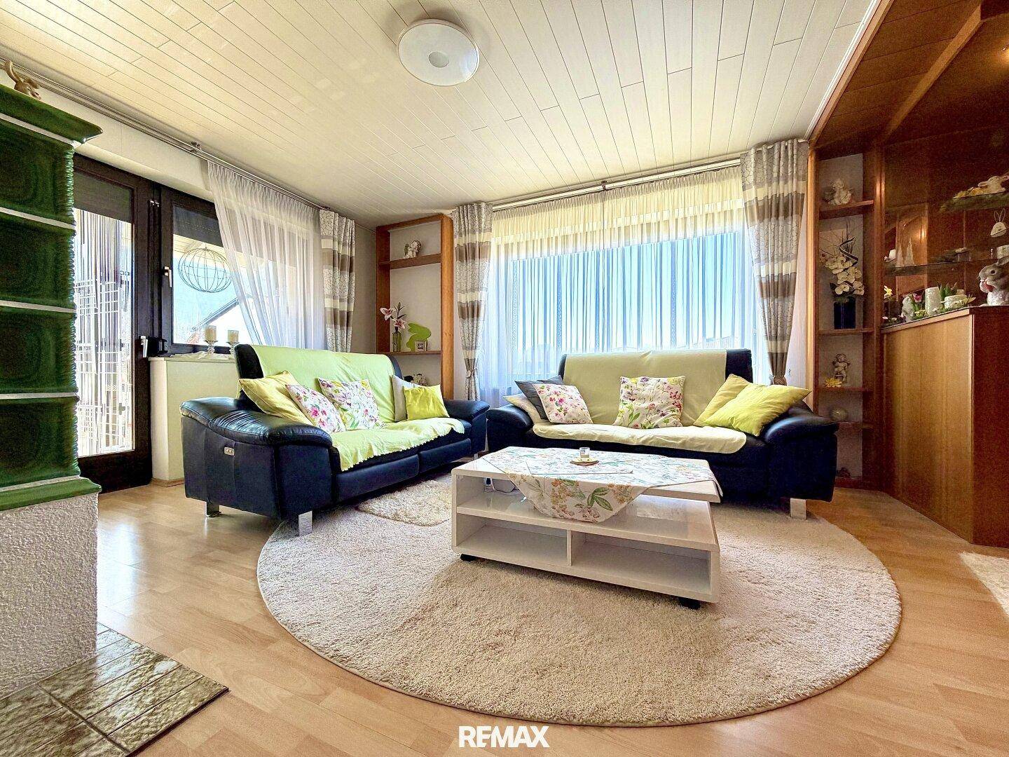 Helles Wohnzimmer mit dunklen Sofas, Laminatboden und einem traditionellen Kachelofen.