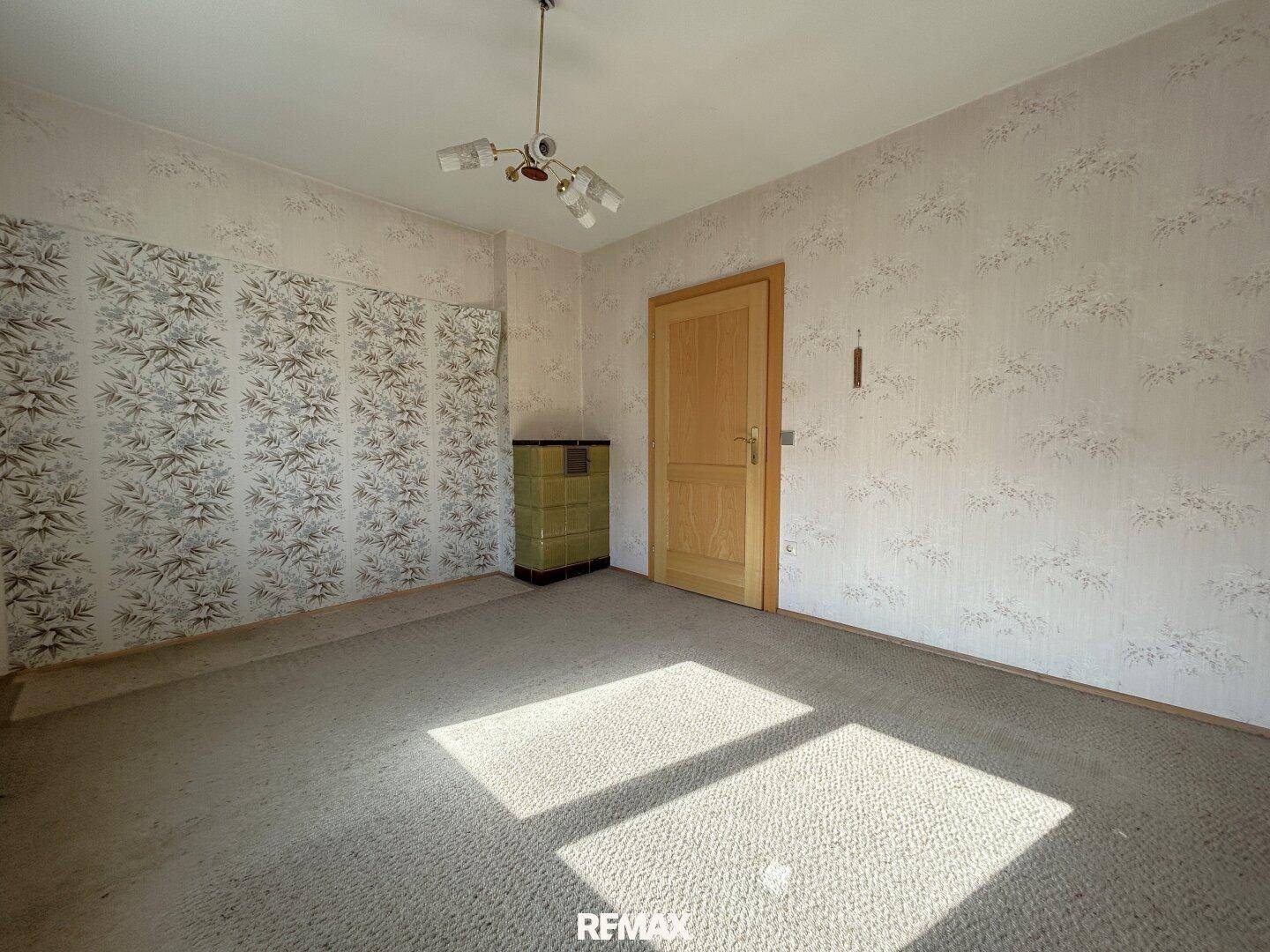 Wohnzimmer mit Teppichboden, Tapeten und einem traditionellen Kachelofen.