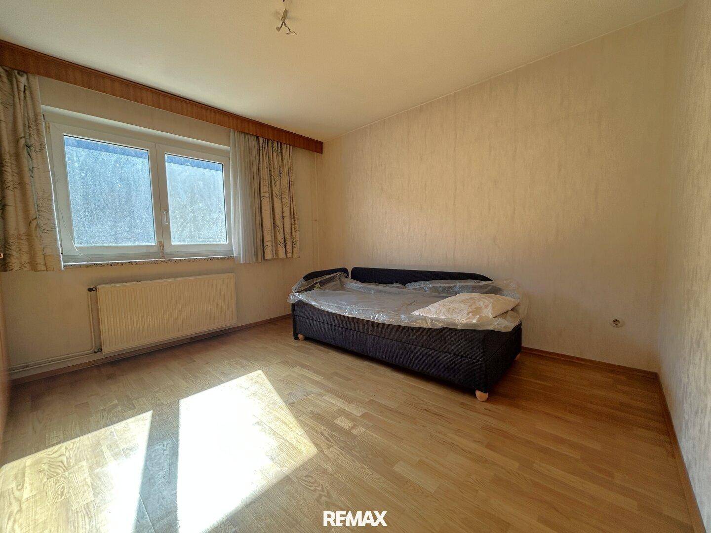 Schlafzimmer mit Holzboden, Tapeten und einem Fenster, das viel Licht hereinlässt.