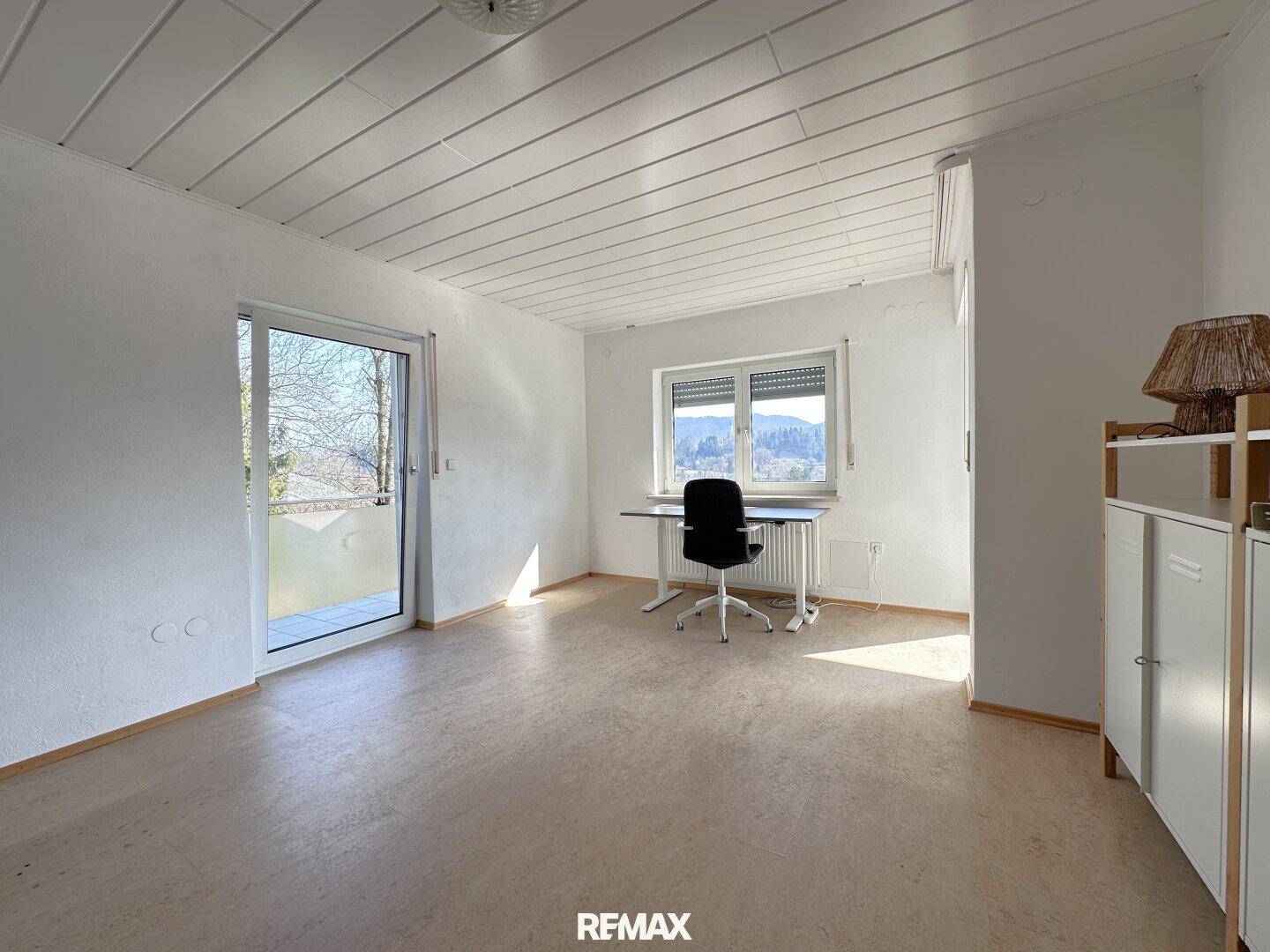 Heller Arbeitsbereich mit Fenster, Blick ins Grüne und einfacher Einrichtung in einem geräumigen Zimmer.
