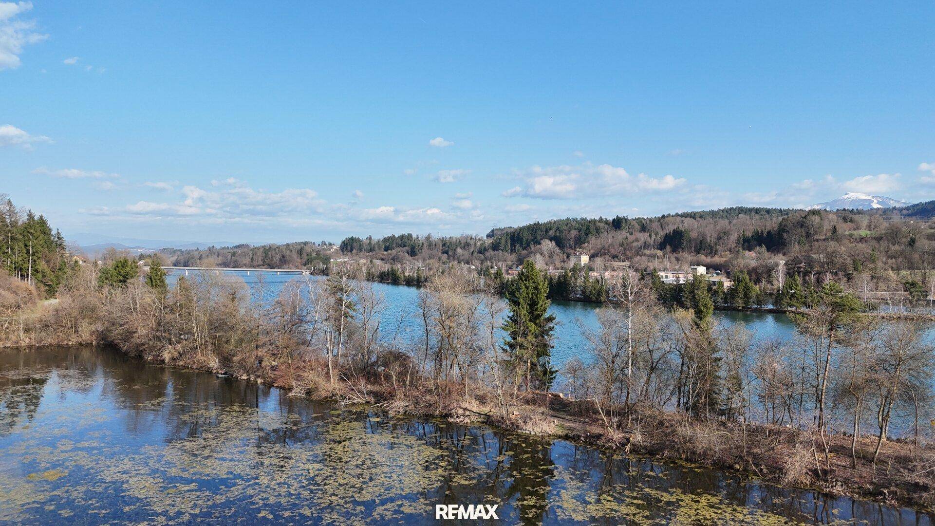 Malerische Seenlandschaft mit bewaldeten Ufern unter einem strahlend blauen Himmel und Bergen im Hintergrund.