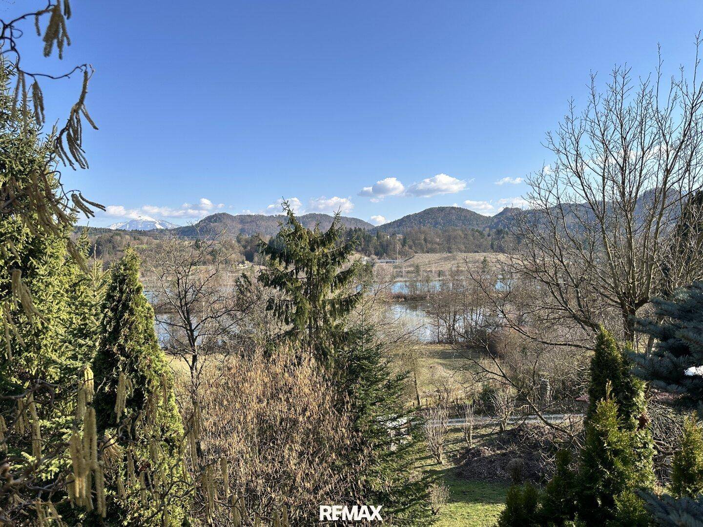 Weitläufiger Ausblick auf die Landschaft mit See, Bergen und bewaldeten Hügeln unter blauem Himmel.