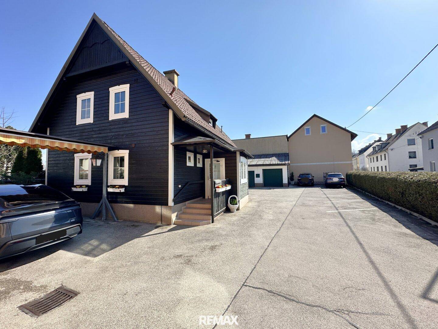 Dunkles Holzhaus mit weißem Giebel und großem Parkplatzbereich vor einem weiteren Gebäude.