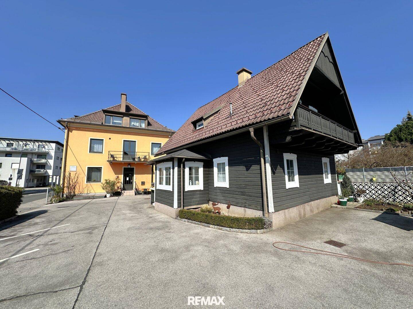 Ansicht eines dunklen Holzhauses und eines orangefarbenen Gebäudes mit großem Parkplatzbereich.