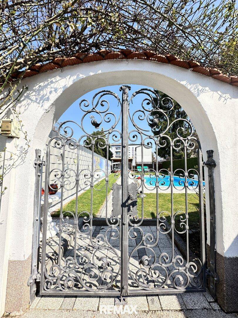 Ein kunstvoll geschmiedetes Tor führt zu einem Gartenbereich mit einem sichtbaren Schwimmbad.