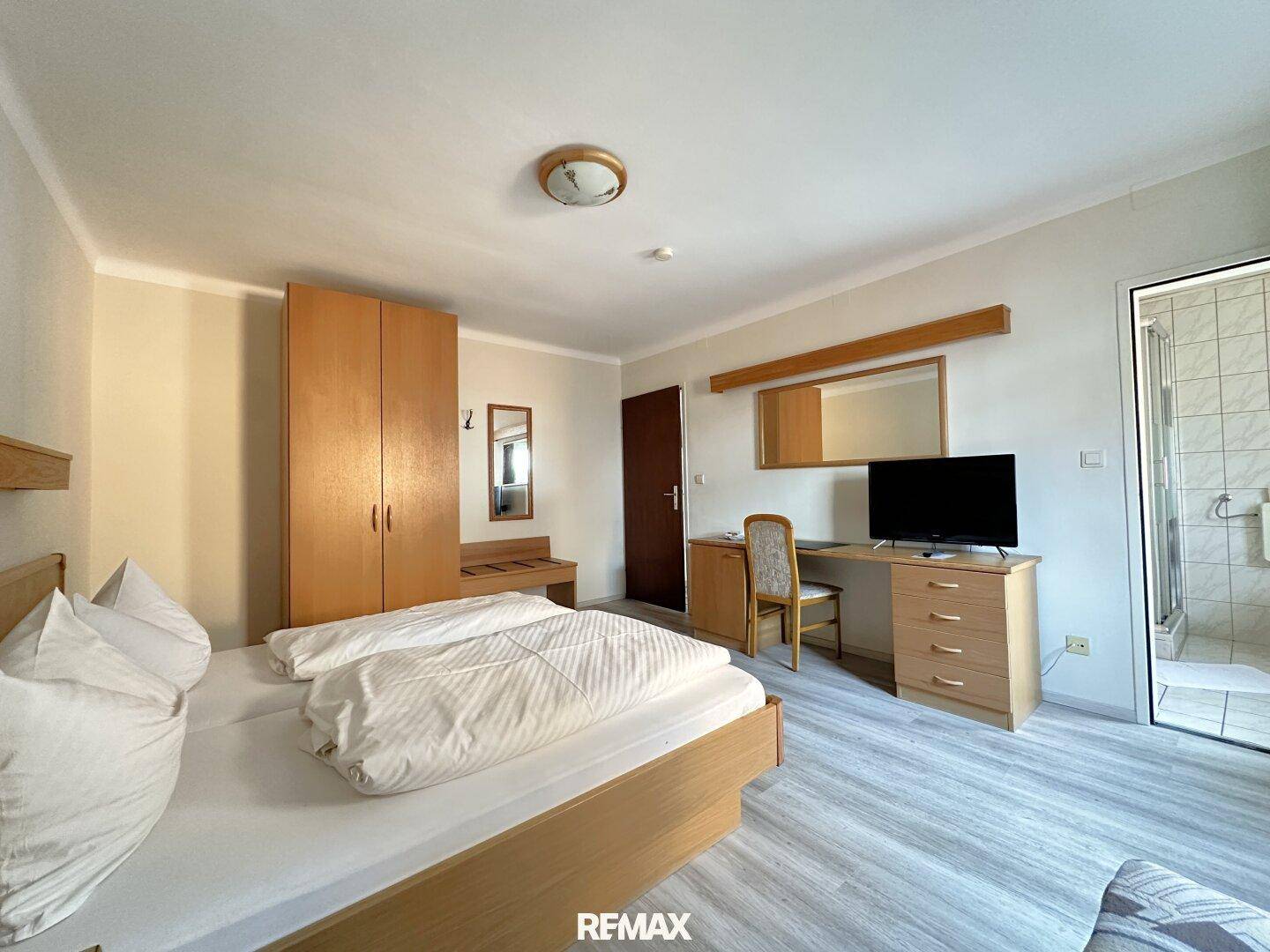 Schlafzimmer mit Doppelbett, Holzschrank, Schreibtisch und direktem Zugang zu einem Badezimmer mit Dusche.