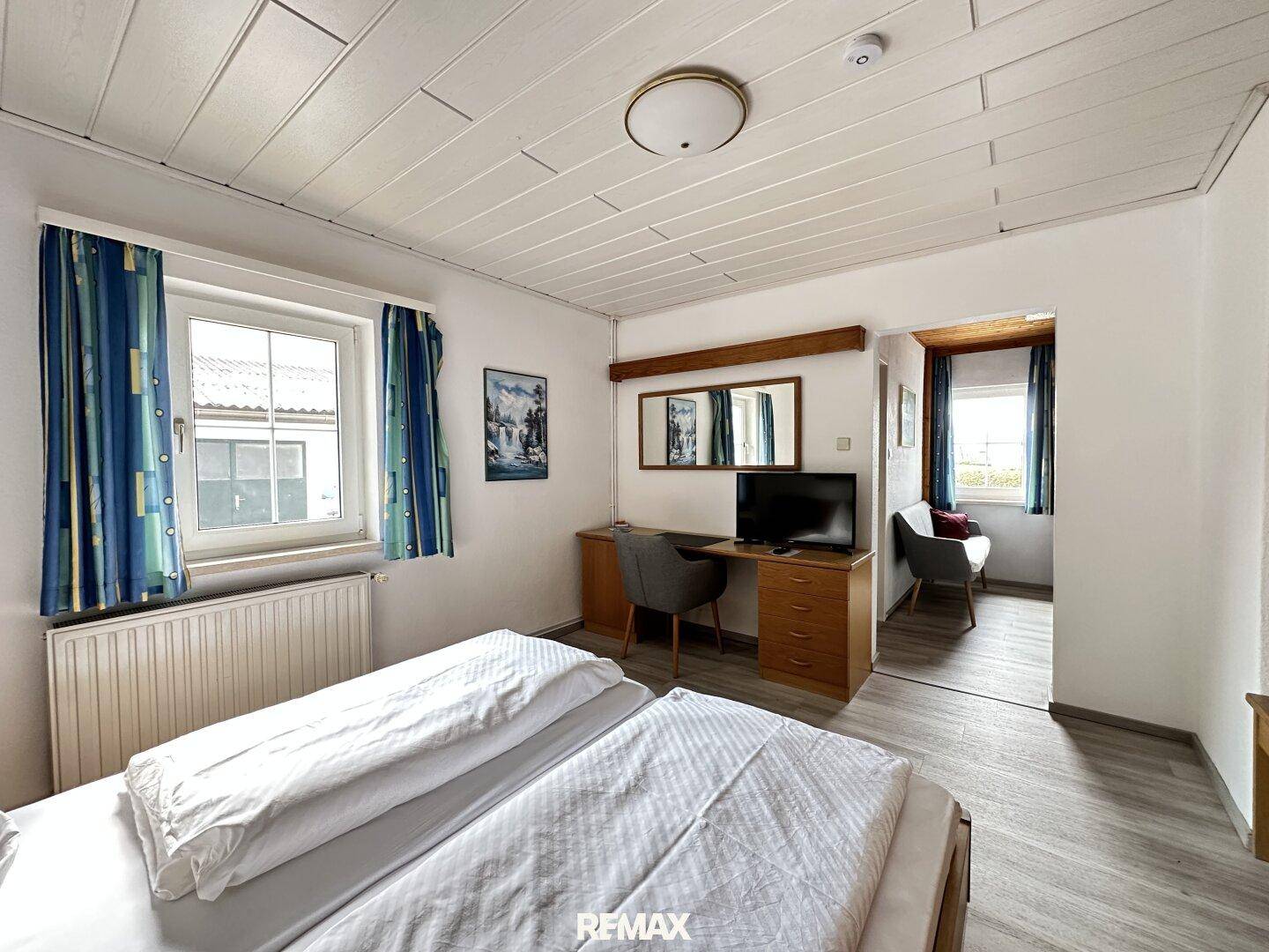 Helles Schlafzimmer mit Doppelbett, Schreibtisch, Fernseher und Zugang zu einem kleinen Sitzbereich.