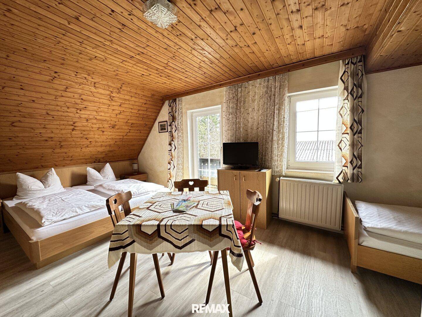 Schlafzimmer mit zwei Betten, Esstisch, Fernseher und einer warmen Holzdecke mit Dachschräge.