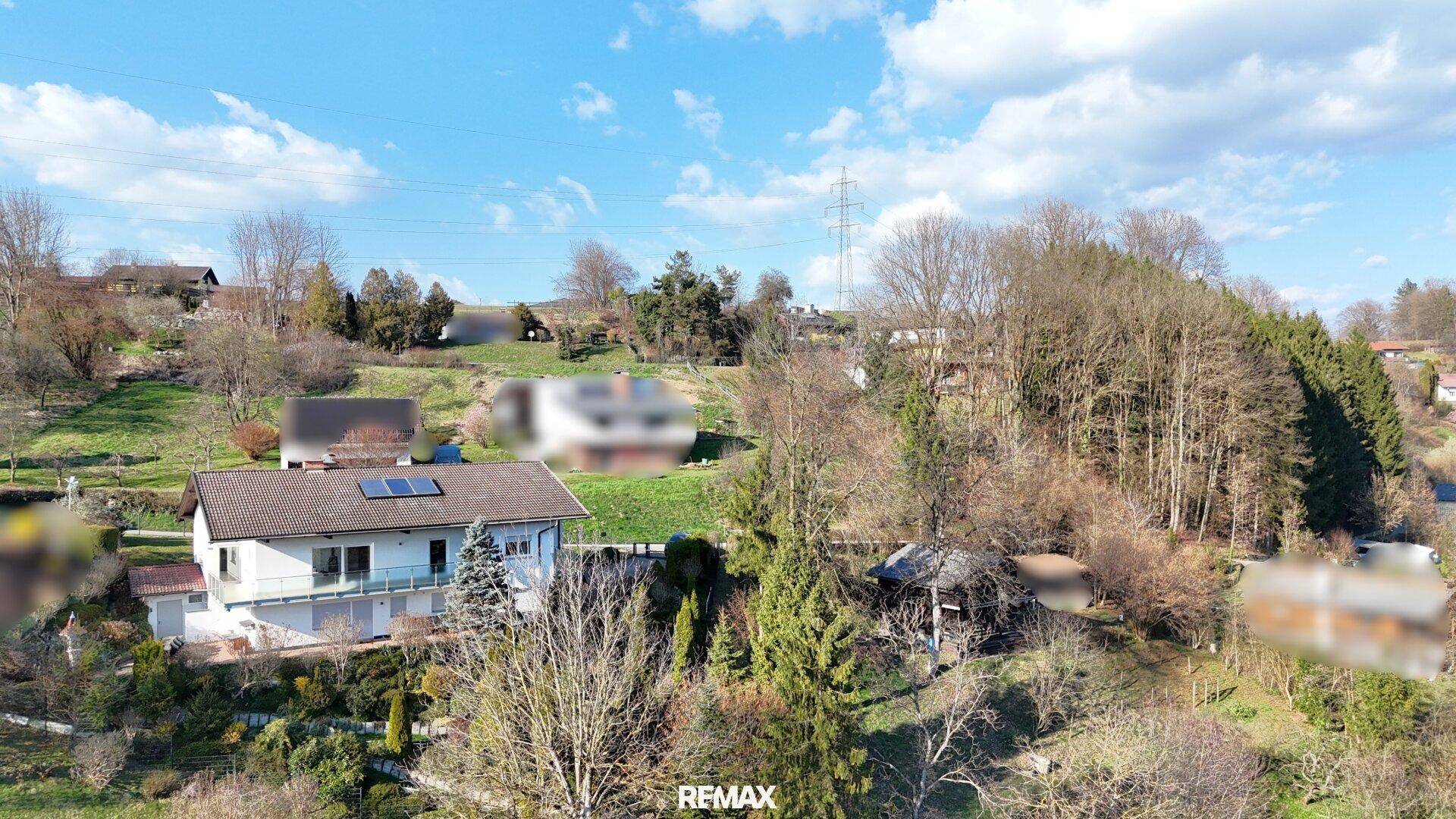 Weitläufige Ansicht des Hauses in einer hügeligen, grünen Landschaft mit weiteren Gebäuden und Stromleitungen.