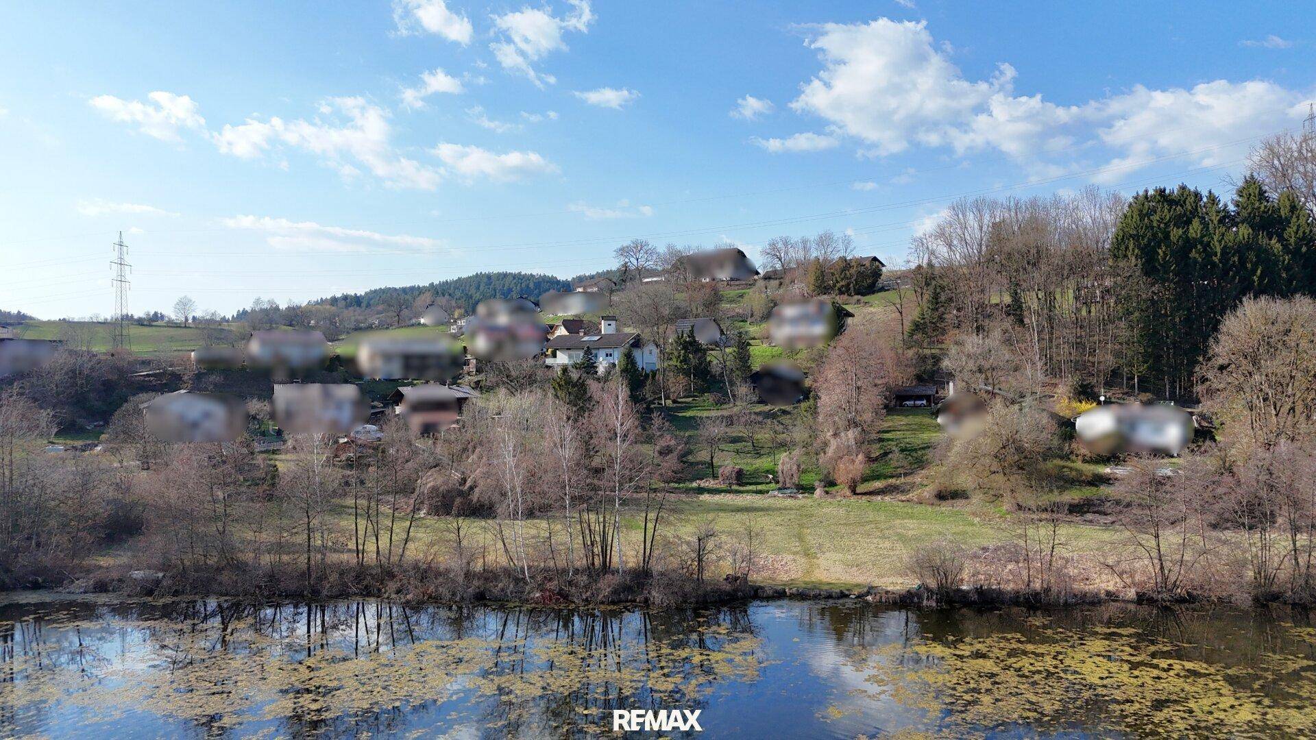 Ruhige Seenlandschaft mit bewaldeten Hügeln und vereinzelten Häusern im Hintergrund unter blauem Himmel.