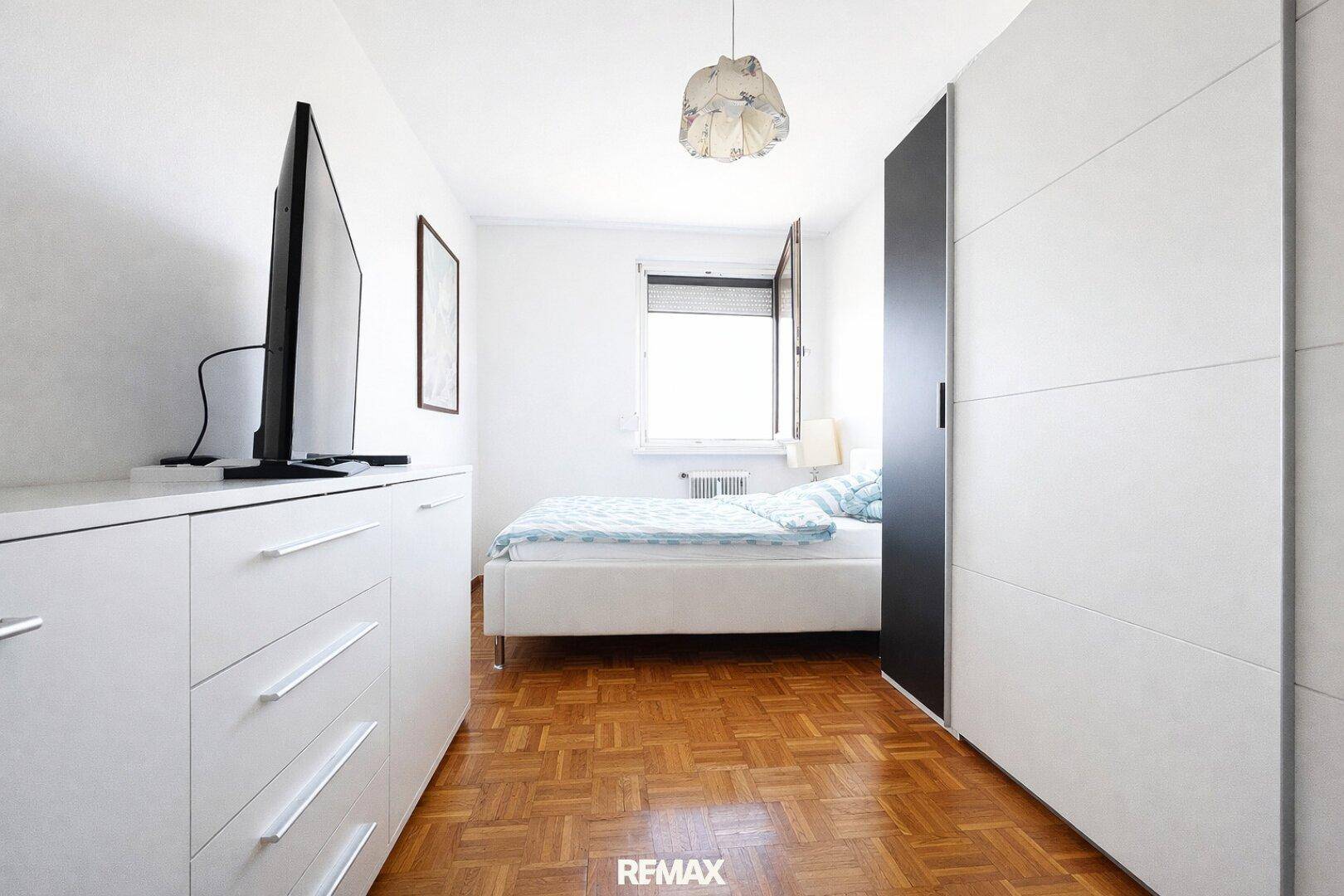 Schlichtes Schlafzimmer mit Parkettboden, weißem Bett und Kleiderschrank.