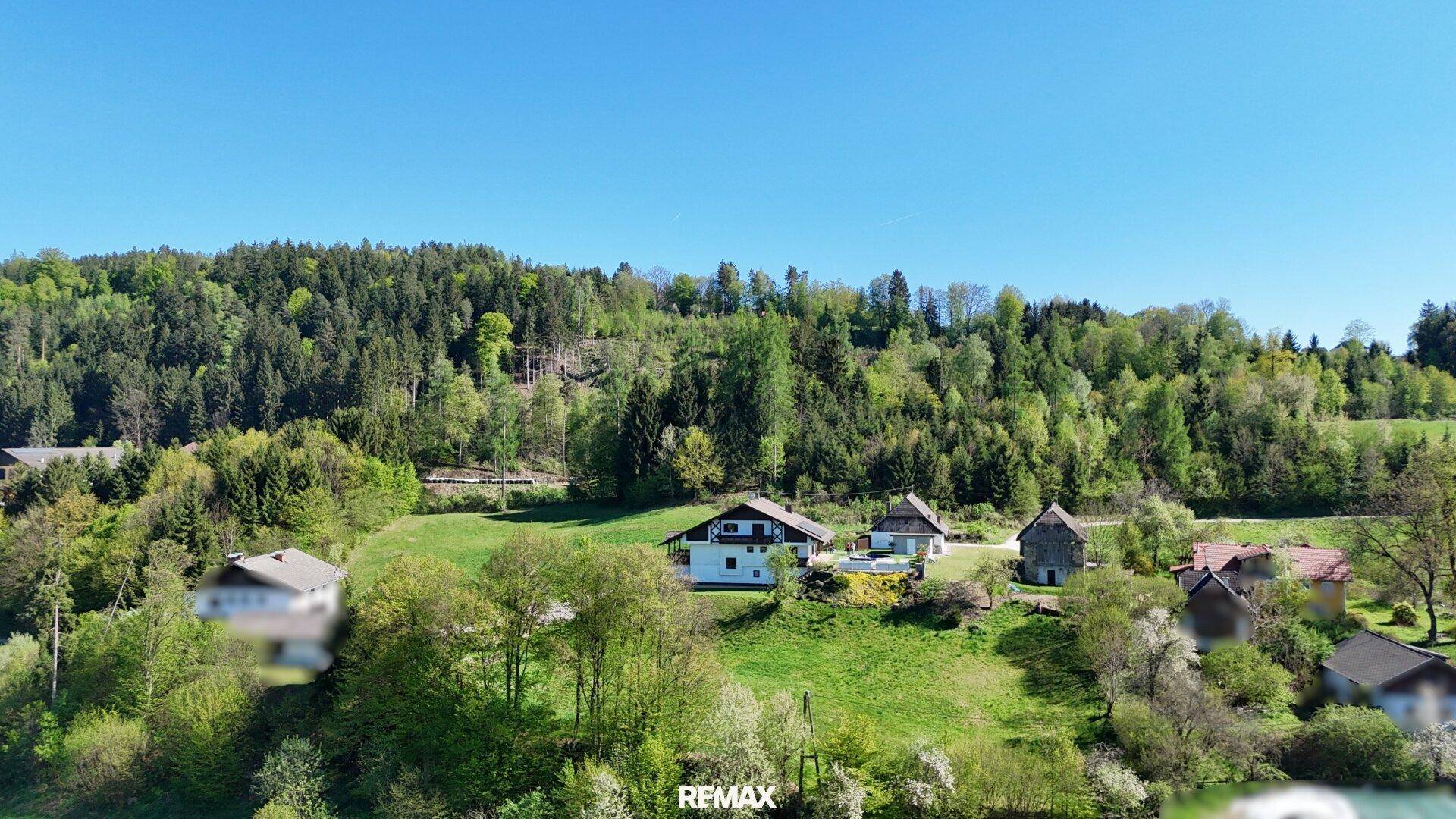 Luftaufnahme einer ländlichen Siedlung mit mehreren Häusern, eingebettet in eine hügelige, bewaldete Landschaft.