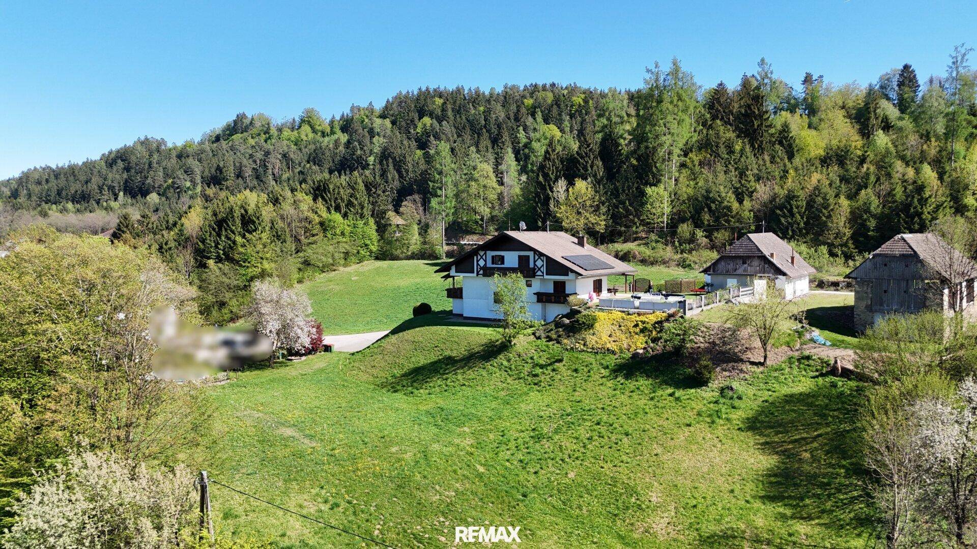 Luftaufnahme eines Anwesens mit Haupthaus, Nebengebäude und Scheune auf einem grünen Hügel vor Waldkulisse.