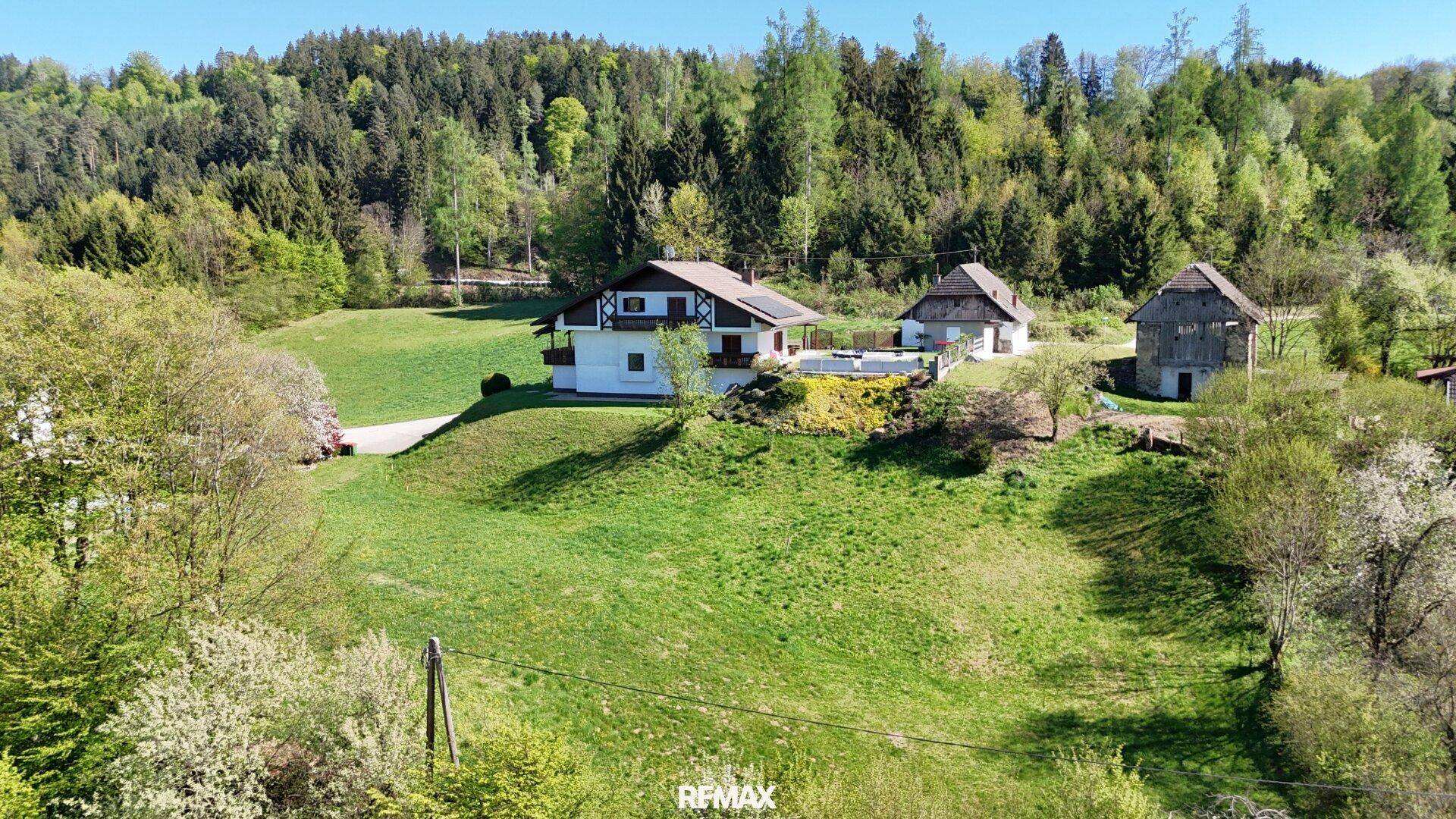 Luftaufnahme eines Anwesens mit Haupthaus, Nebengebäude und Scheune auf einem grünen Hügel vor Waldkulisse.