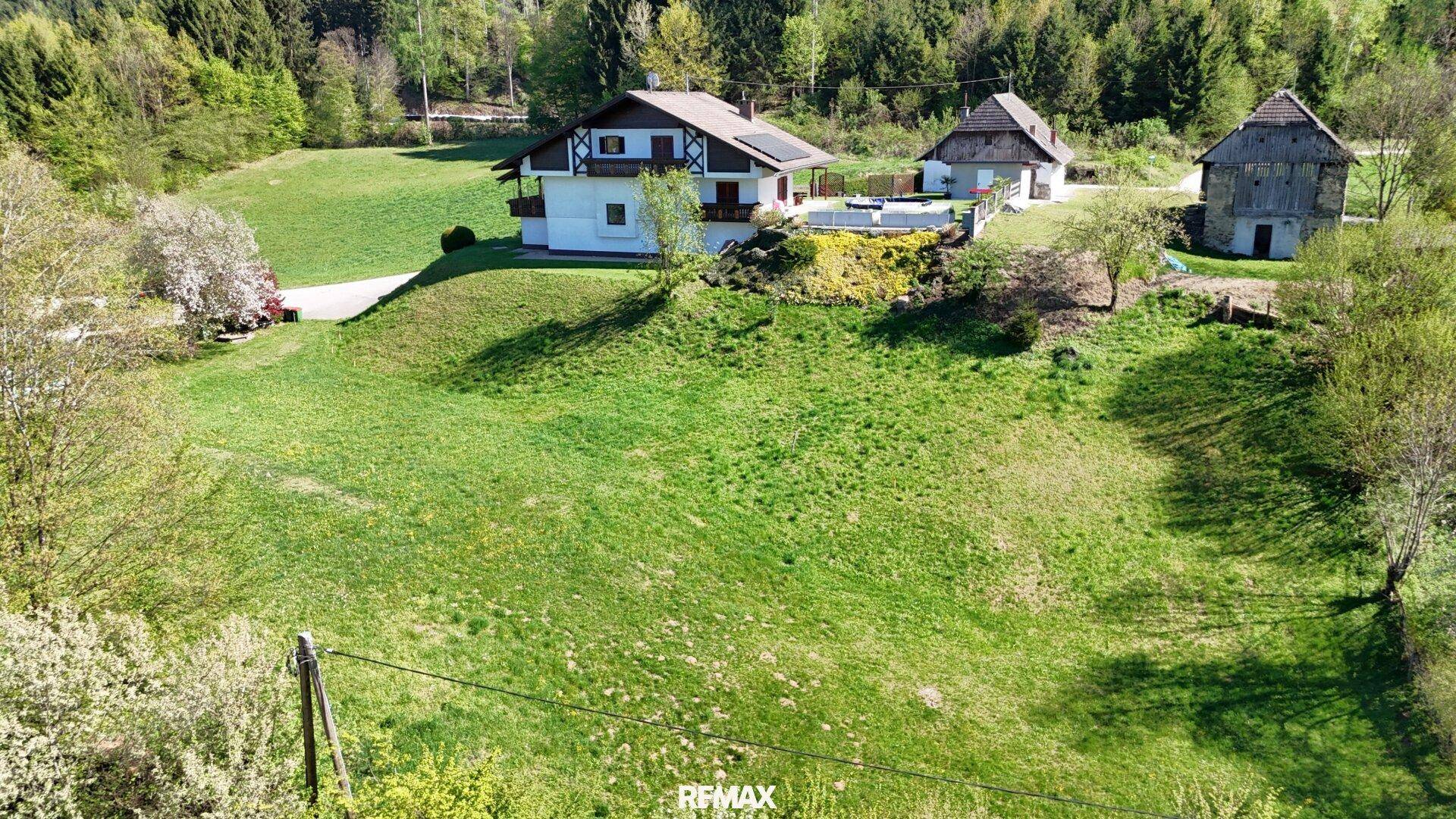 Luftaufnahme eines Anwesens mit Haupthaus, Nebengebäude und Scheune auf einem grünen Hügel vor Waldkulisse.