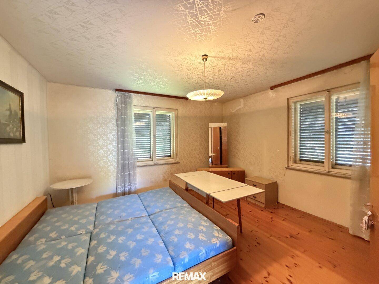 Schlafzimmer mit Holzfußboden, zwei Fenstern und einfacher Einrichtung.