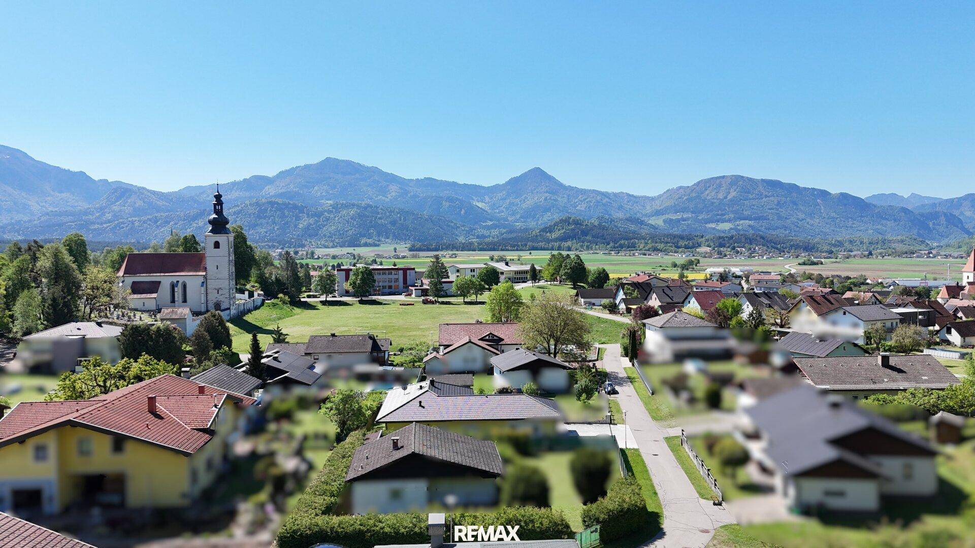 Weitläufiger Blick über eine ländliche Siedlung mit Kirche und majestätischer Bergkulisse im Hintergrund.