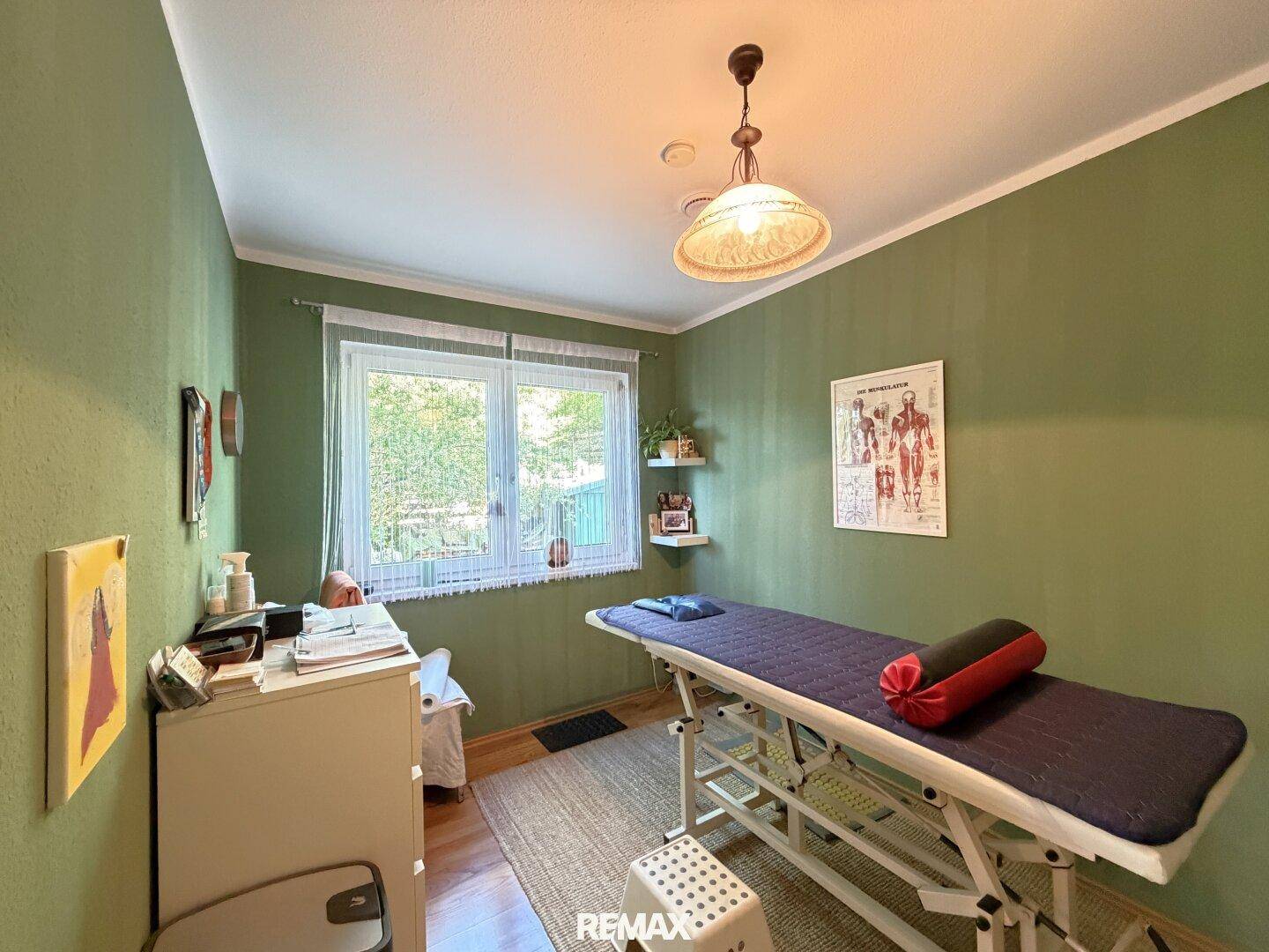 Grüner Raum, eingerichtet als Behandlungs- oder Arbeitszimmer mit Fenster und Holzboden.