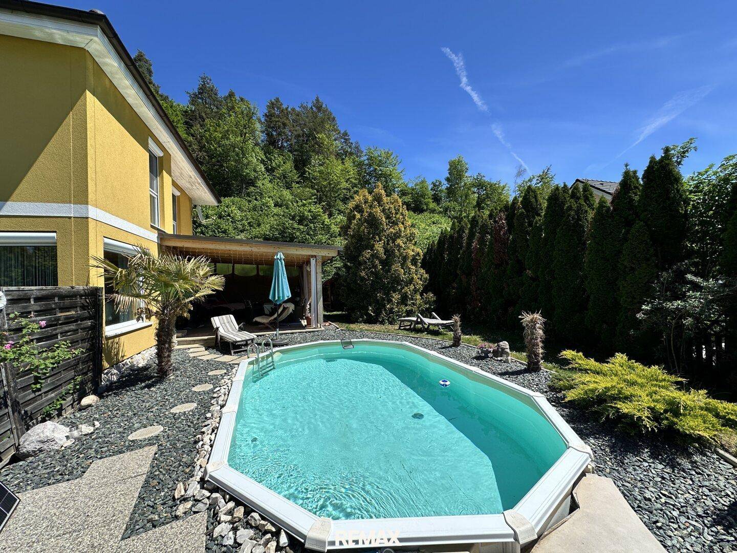 Außenpoolbereich mit Liegestühlen, Palmen und überdachter Terrasse, umgeben von viel Grün.