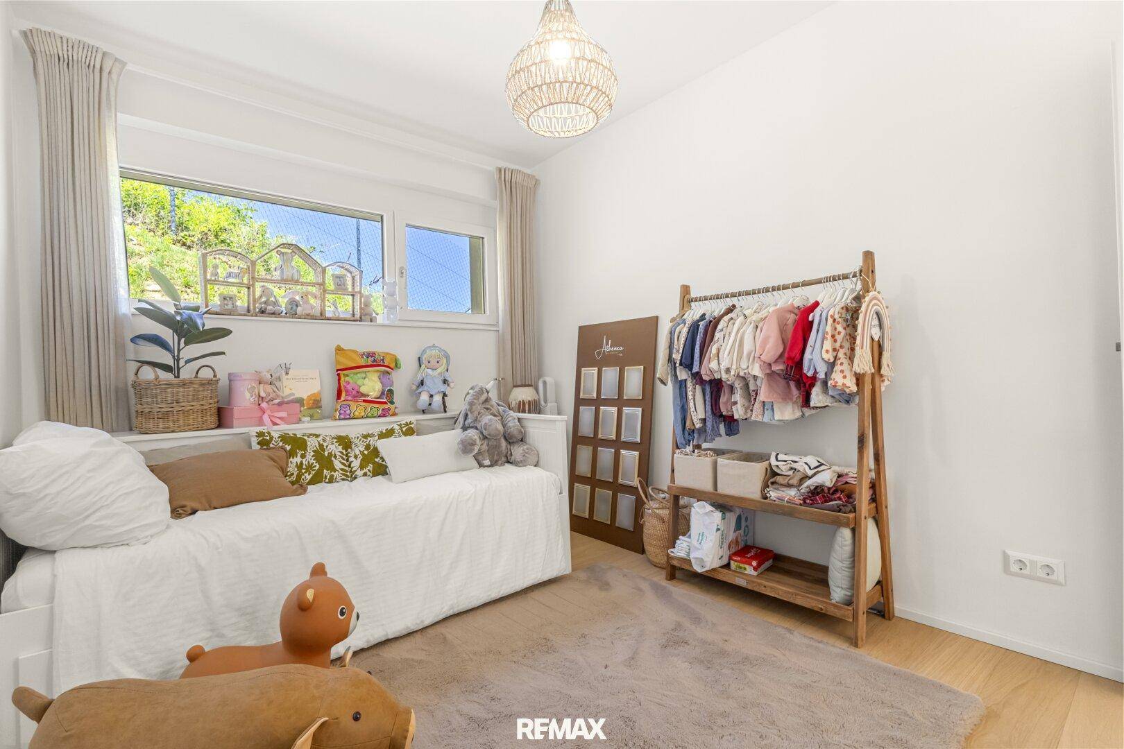 Helles Kinderzimmer mit Einzelbett, Kleiderstange und Blick ins Grüne durch das Fenster.