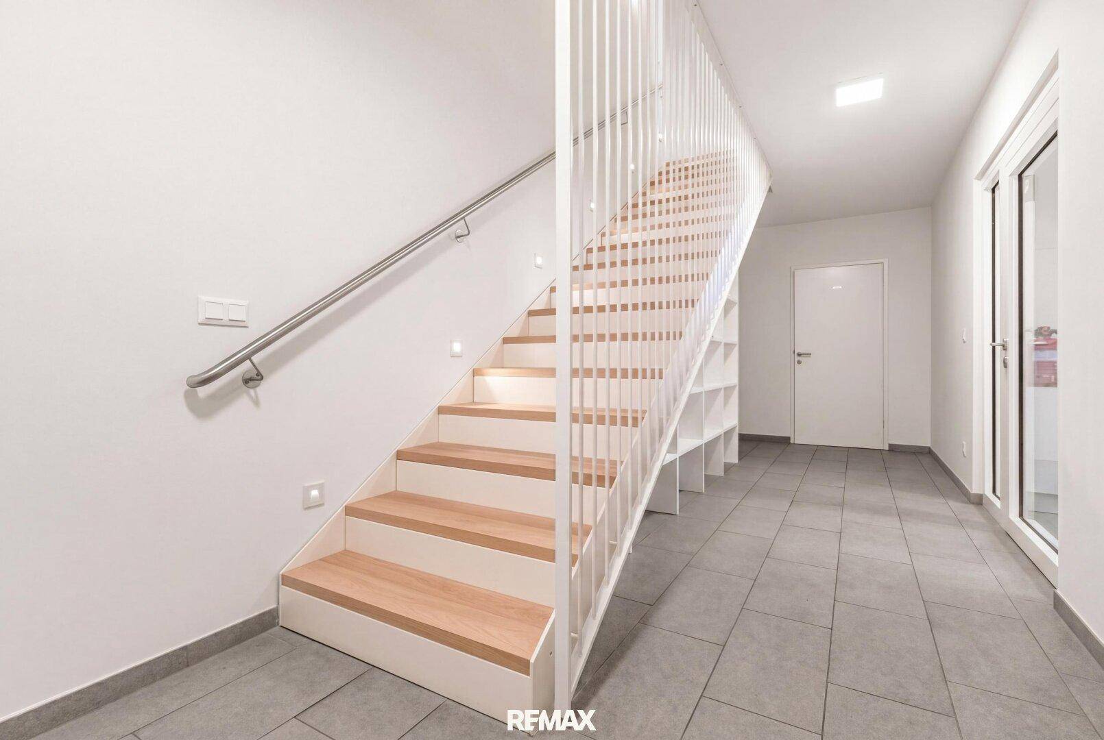 Heller Flur mit einer modernen Holztreppe, integrierten Regalen und Fliesenboden.